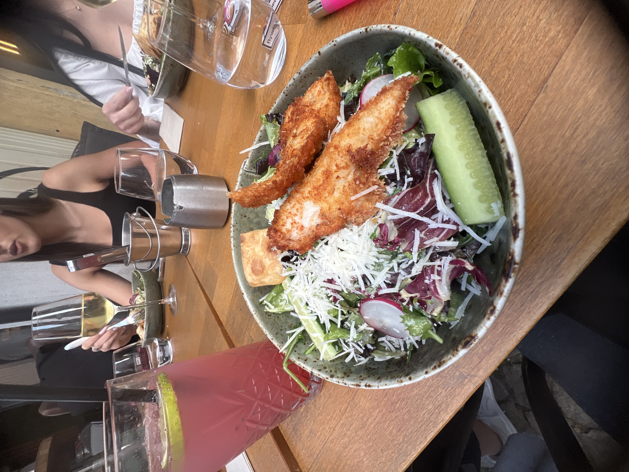 A high-angle, indoor shot depicts a wooden table with a bowl of salad, drinks, and dining companions. The salad contains lettuce, fried fish, shredded cheese, radishes, and sliced cucumber. A glass of pink lemonade with a lime wedge and a glass of white wine are visible. A person in a black tank top sits to the left of the table, and another person in a white top is partially visible at the top left. Silverware and a small metal container are also present. The location is likely a restaurant in the city of Plovdiv, Bulgaria.