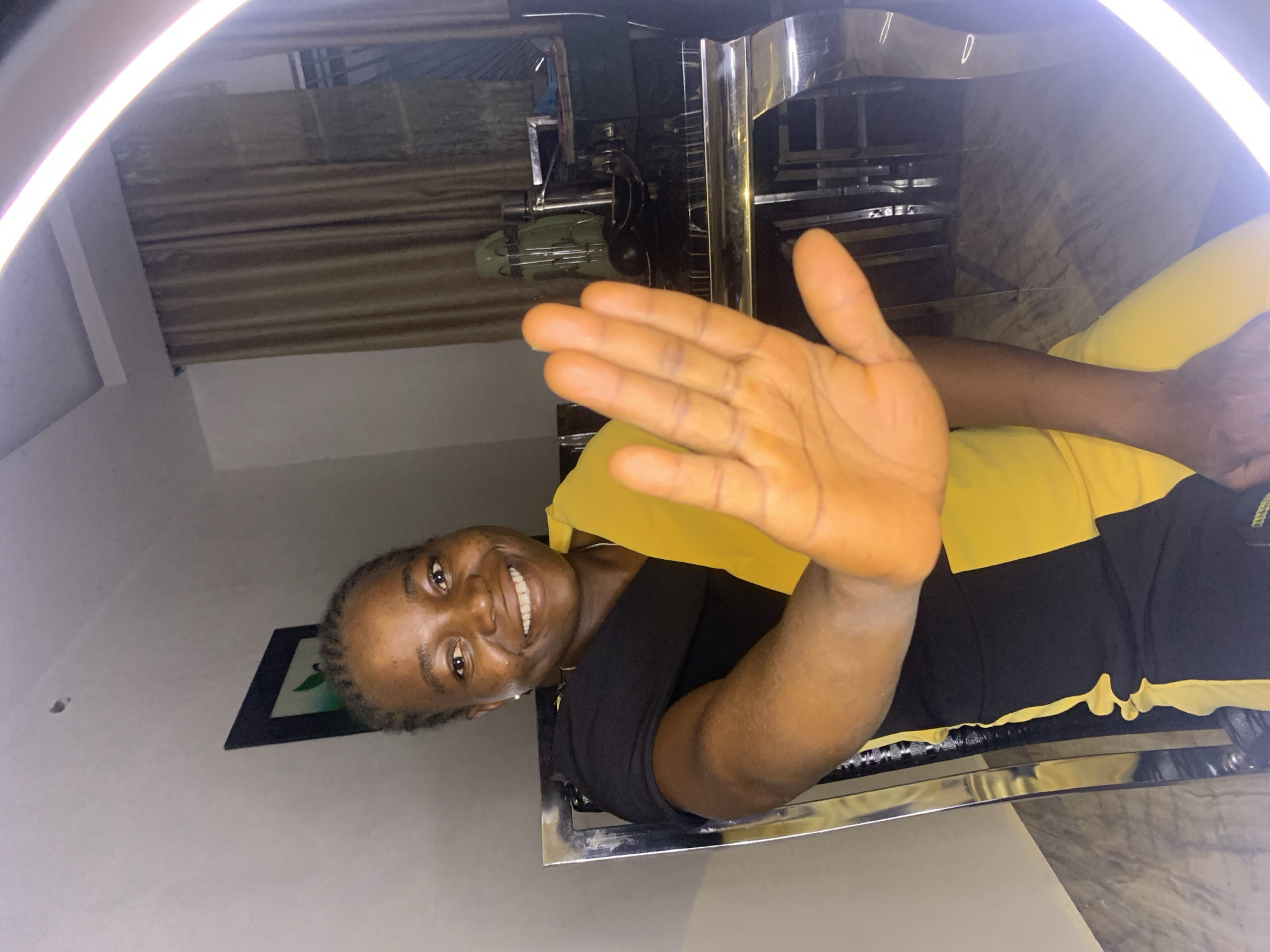 A woman smiles and holds her right hand up in a waving gesture. She wears a black and yellow garment. She appears to be sitting at a metallic structure of sorts. The background includes a wall, a framed picture, a window, and a corrugated metal roof. The location is possibly Jos, Nigeria.