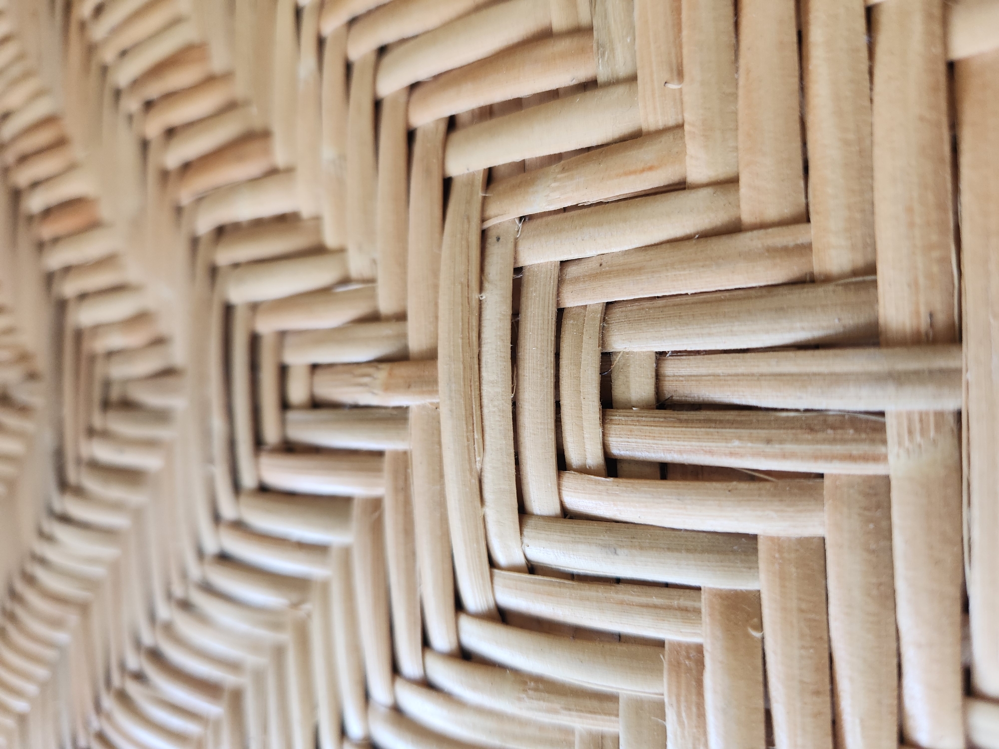 This is a close-up, high-angle shot of a woven cane or rattan material. The pattern is an over-under weave, with thin, light-colored strands interlacing to create a textured surface. The strands appear to be made of natural material, likely wood or bamboo, and exhibit variations in color from pale yellow to light brown, with some darker marks and imperfections. The lighting is diffused, creating soft shadows and highlighting the three-dimensional quality of the weave. The perspective is shallow, with the foreground in sharp focus and the background becoming progressively blurred, suggesting a close-up view of an object such as a chair seat or basket. The material is consistent and uniform across the frame. There are no people, actions, or interactions depicted. The scene is devoid of any contextual environmental details, such as a specific location within Maitland, United States of America.