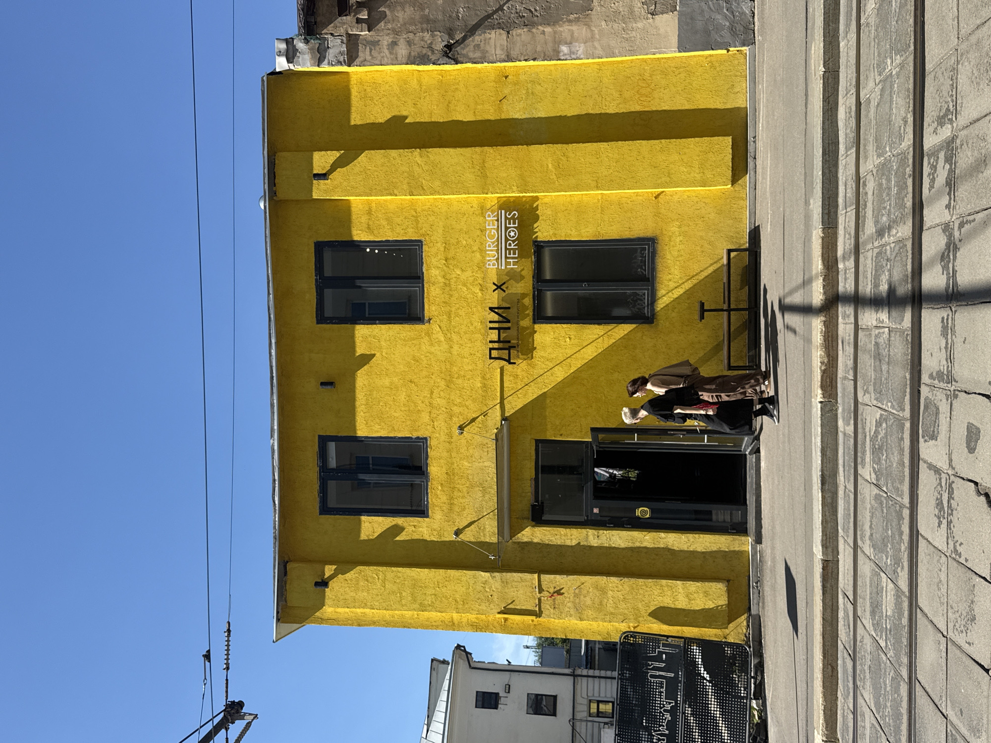 This image depicts the exterior of a bright yellow building located in Moscow, Russia. The building features dark-framed windows and a prominent entrance with an open door. Above the entrance, a sign displays text in Cyrillic and English: "-дни x BURGER HEROES". Two individuals, one with blonde hair wearing a black dress and the other with darker hair wearing a brown outfit, are walking towards the building entrance. To the left of the yellow building, a portion of a white building with windows is visible. In the foreground, tram tracks are laid on a paved street, suggesting an urban environment. Overhead, electrical wires traverse the bright blue sky.