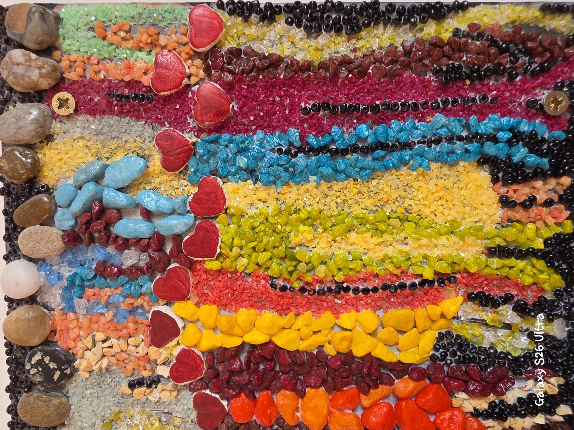 This is a close-up, top-down view of a textured mosaic artwork. The artwork is primarily composed of small, colorful, irregularly shaped stones and beads arranged in vertical stripes and abstract patterns. Dominant colors include red, yellow, orange, blue, green, purple, brown, and black. Several red heart-shaped objects are incorporated into the mosaic. The background appears to be a light-colored, textured surface. A watermark in the bottom left corner reads "Galaxy S26 Ultra". The artwork is not located in a specific city in Plovdiv, Bulgaria; it is an indoor artwork.