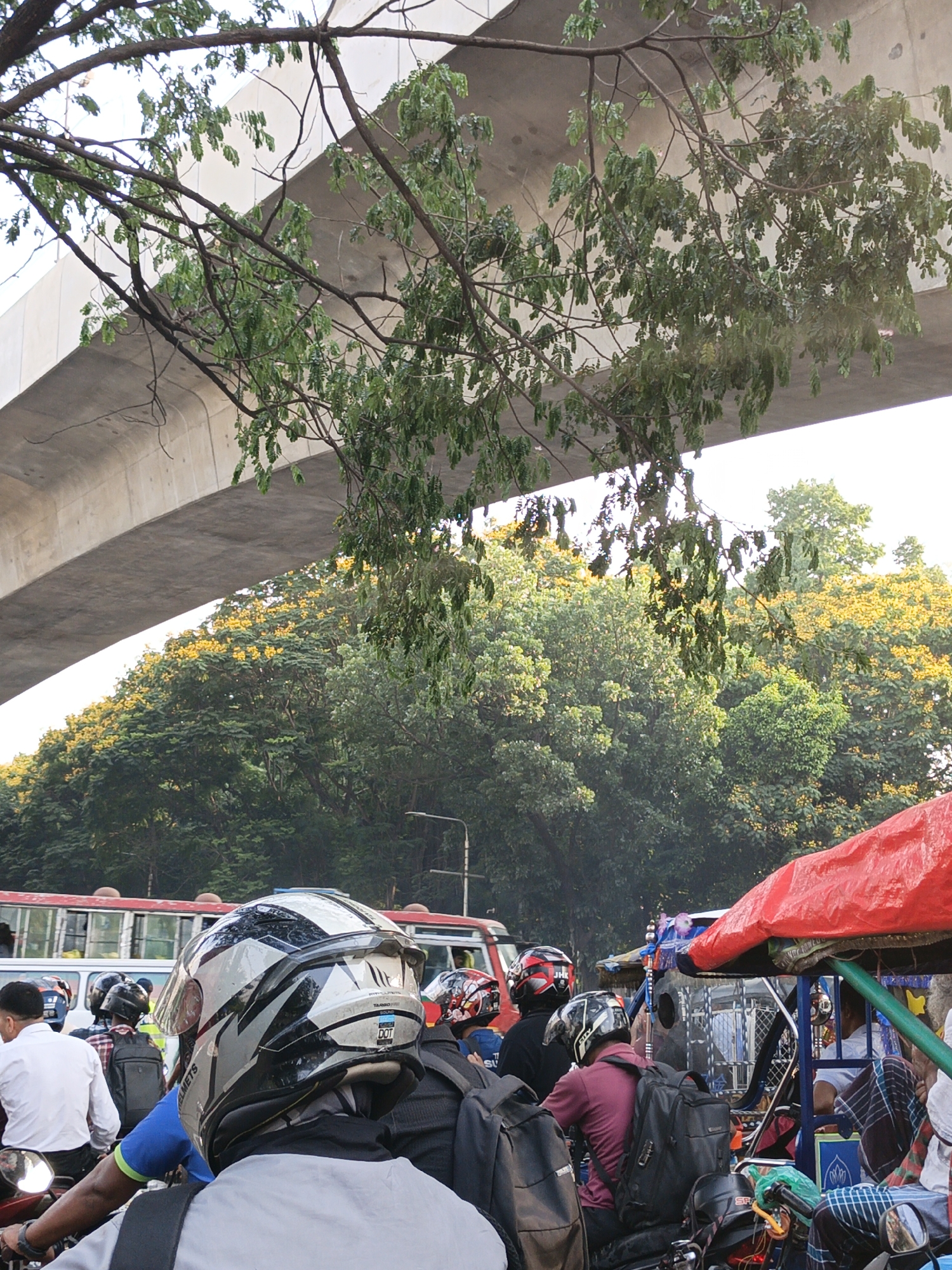 The image shows a busy street scene in an urban area, possibly in Unknown, Unknown. An elevated concrete highway structure looms overhead, partially obscured by tree branches with green leaves. Below the highway, a row of trees with yellow blossoms is visible.
In the foreground, a group of motorcyclists wearing helmets and backpacks are stopped in traffic. Several individuals are wearing red, black and white helmets. In front of the motorcyclists, a bus and a covered tricycle-type vehicle (likely a rickshaw or similar mode of transport) are present. The scene suggests heavy traffic congestion.