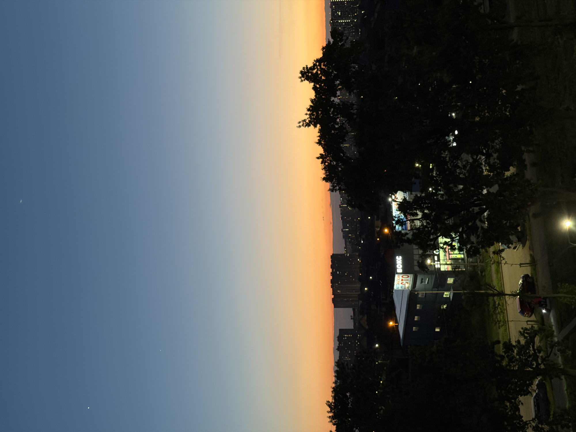 The image depicts a twilight scene over a city, likely Krasnodar, Russia, given the Cyrillic signage. The sky transitions from a deep blue at the top to a vibrant orange and yellow hue near the horizon, indicating either sunrise or sunset. Two celestial bodies, possibly planets or bright stars, are visible in the upper portion of the sky. Silhouetted against the colorful sky are distant apartment buildings and urban structures. In the foreground, a dark tree canopy obscures parts of the view. Below the horizon, streetlights illuminate a street and a building with Cyrillic text visible, including "АВТОМАГАЗИН" and "СТО" and "БОКС", suggesting a retail or service area. Cars are parked or driving on the street. The overall atmosphere is serene and urban.