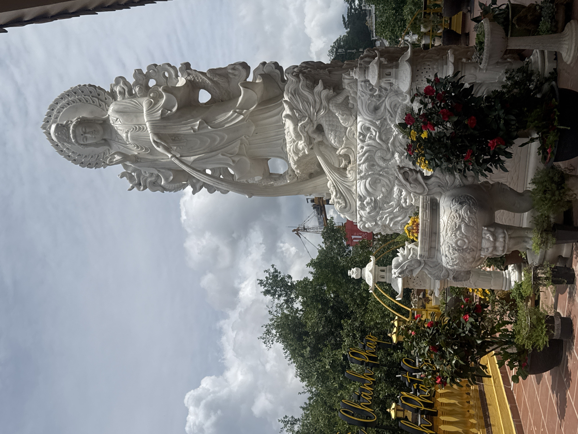 The image depicts a large white marble statue of a female deity, likely Quan Am (Goddess of Mercy), in a temple courtyard in Da Lat, Vietnam. The statue stands on an ornate base adorned with dragons and swirling patterns. She is depicted with flowing robes and is holding a vase from which water appears to be pouring. Flanking the statue are smaller white marble pagodas and large stone incense burners. In the foreground, potted plants with red and yellow flowers are arranged. A bright yellow sign with Vietnamese text is visible to the left, indicating this is likely a Buddhist temple or a tourist attraction. The background shows lush green trees, a cloudy sky, and a construction crane in the distance, suggesting urban development in the area. The overall scene conveys a sense of serenity and cultural significance, set within a tropical environment.