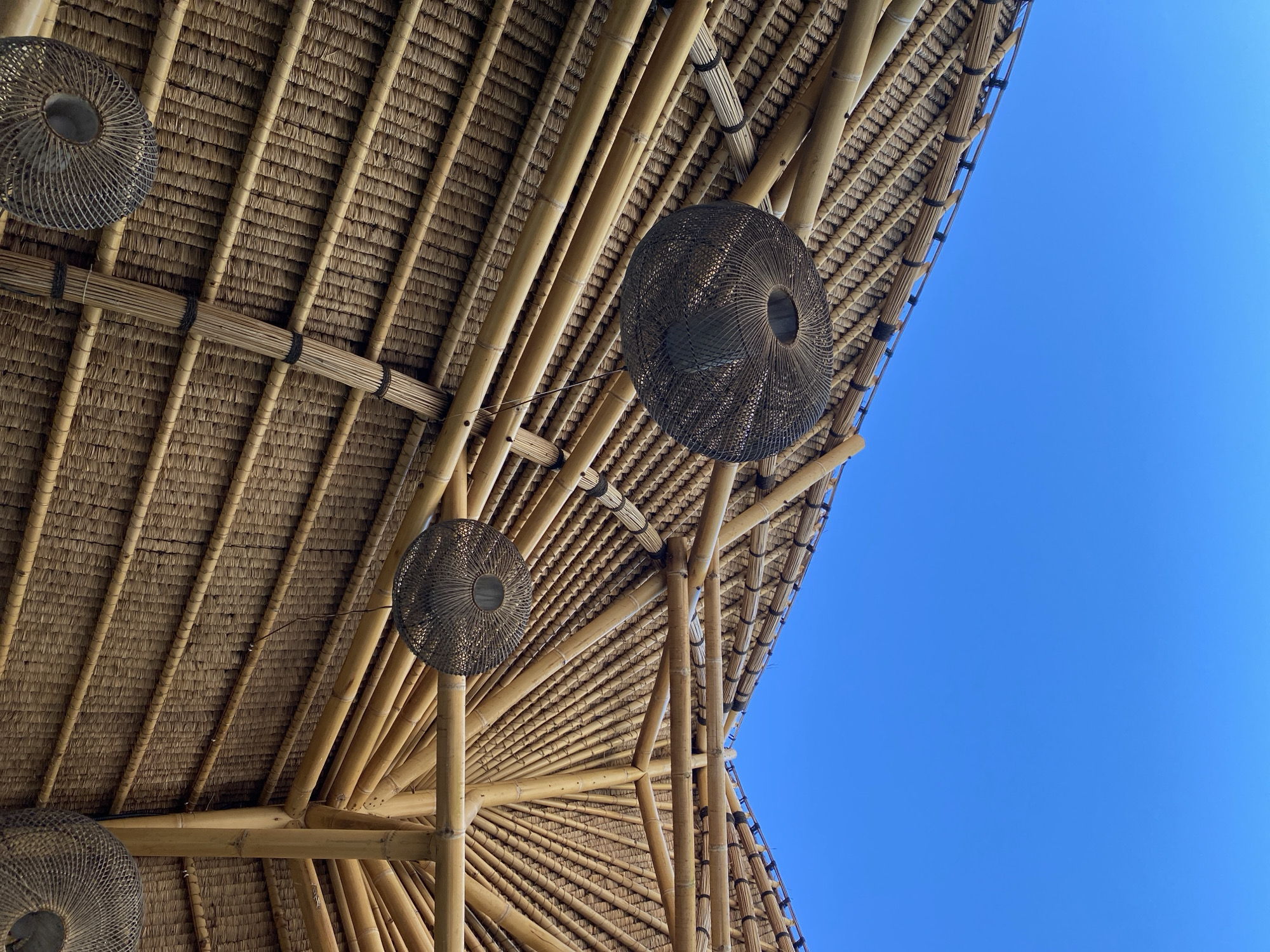 The image depicts the underside of a thatched roof structure in Kuta Selatan, Indonesia. The roof is constructed of bamboo beams and woven straw, arranged in a dense pattern. Several spherical, woven lampshades hang from the roof structure. The background consists of a clear, bright blue sky.