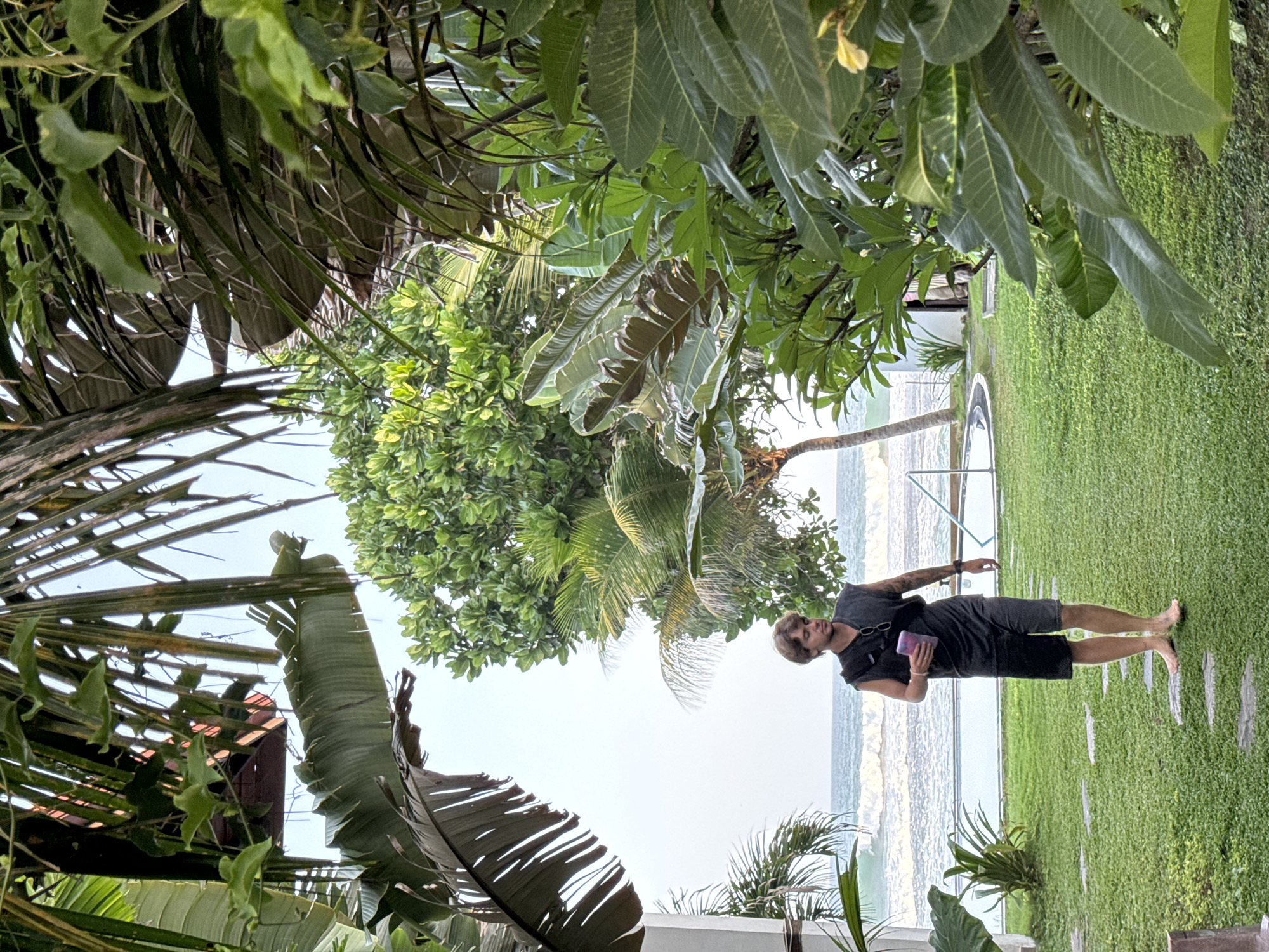 A young Caucasian male with brown hair walks on a stepping stone path across a neatly manicured green lawn. He is wearing a sleeveless dark grey t-shirt and dark grey shorts. He has a dark bag slung across his chest and is holding a purple tumbler in his right hand. Behind him, a blue swimming pool is visible with the ocean and waves in the background. Tropical foliage frames the scene, with palm leaves prominent in the foreground and mid-ground. The sky is a pale blue. The location is Goviyapana, Sri Lanka.
