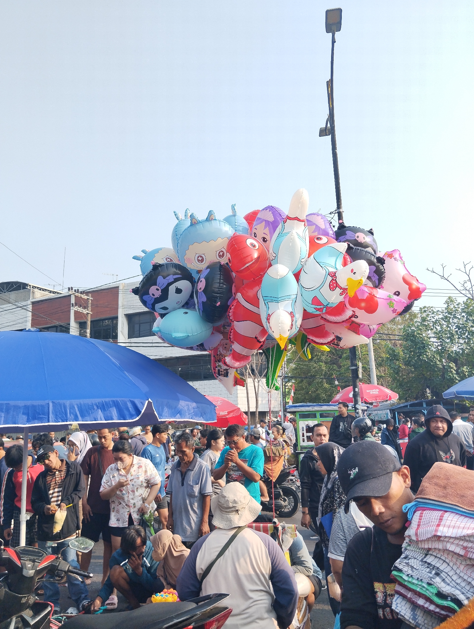 This image depicts a bustling outdoor market scene, likely in Surabaya, Indonesia, characterized by a lively crowd and a variety of goods. Dominating the upper portion of the frame is a large bunch of colorful, character-shaped balloons, including those resembling cartoon animals and figures, tied to a lamppost. Below the balloons, a vibrant blue umbrella shades a section of the market, hinting at vendor stalls. The ground is filled with a diverse group of people, many appearing to be shoppers and vendors engaged in various activities. Some individuals are inspecting goods, others are in conversation, and some are simply navigating the crowded space. Motorcycles are visible parked in the foreground and midground, suggesting transportation within the market area. The overall atmosphere is one of active commerce and community interaction, typical of a local market in a densely populated urban environment. The presence of street vendors, stalls, and a congregation of people indicates a vibrant marketplace.