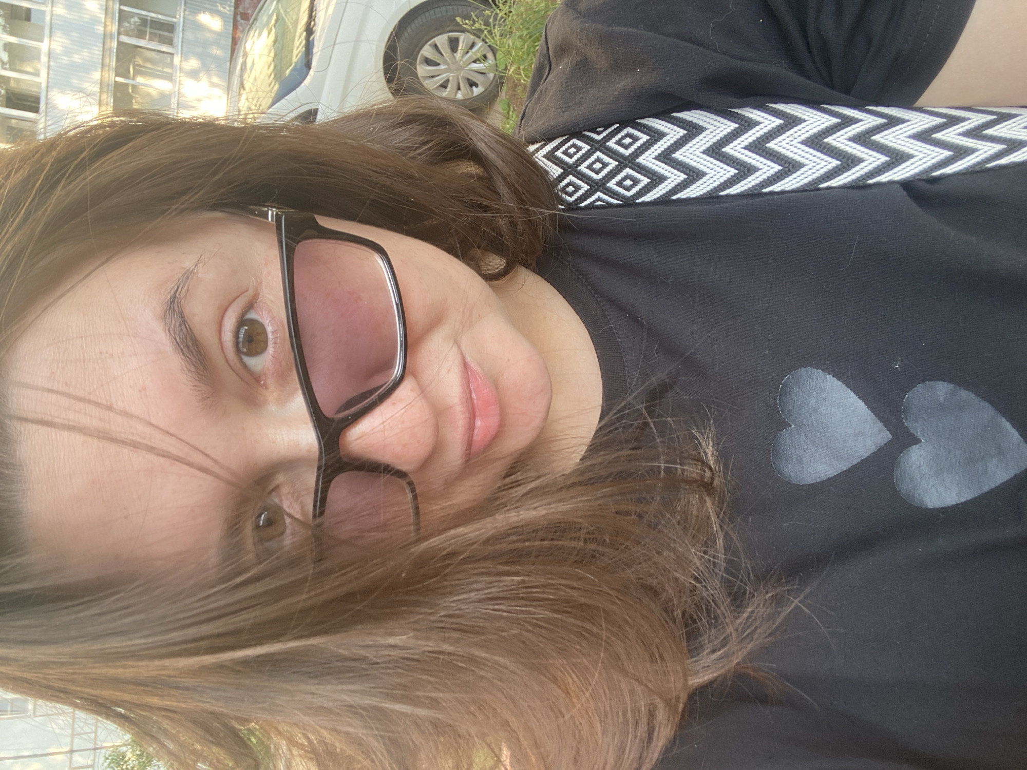 The original image is a selfie of a woman taken outdoors. She has brown hair and is wearing sunglasses and a black t-shirt with two grey hearts printed on it. A patterned strap of a bag is visible over her shoulder. In the background, there is a white car parked and a building with balconies. This scene is located in Chaikovsky, Russia.