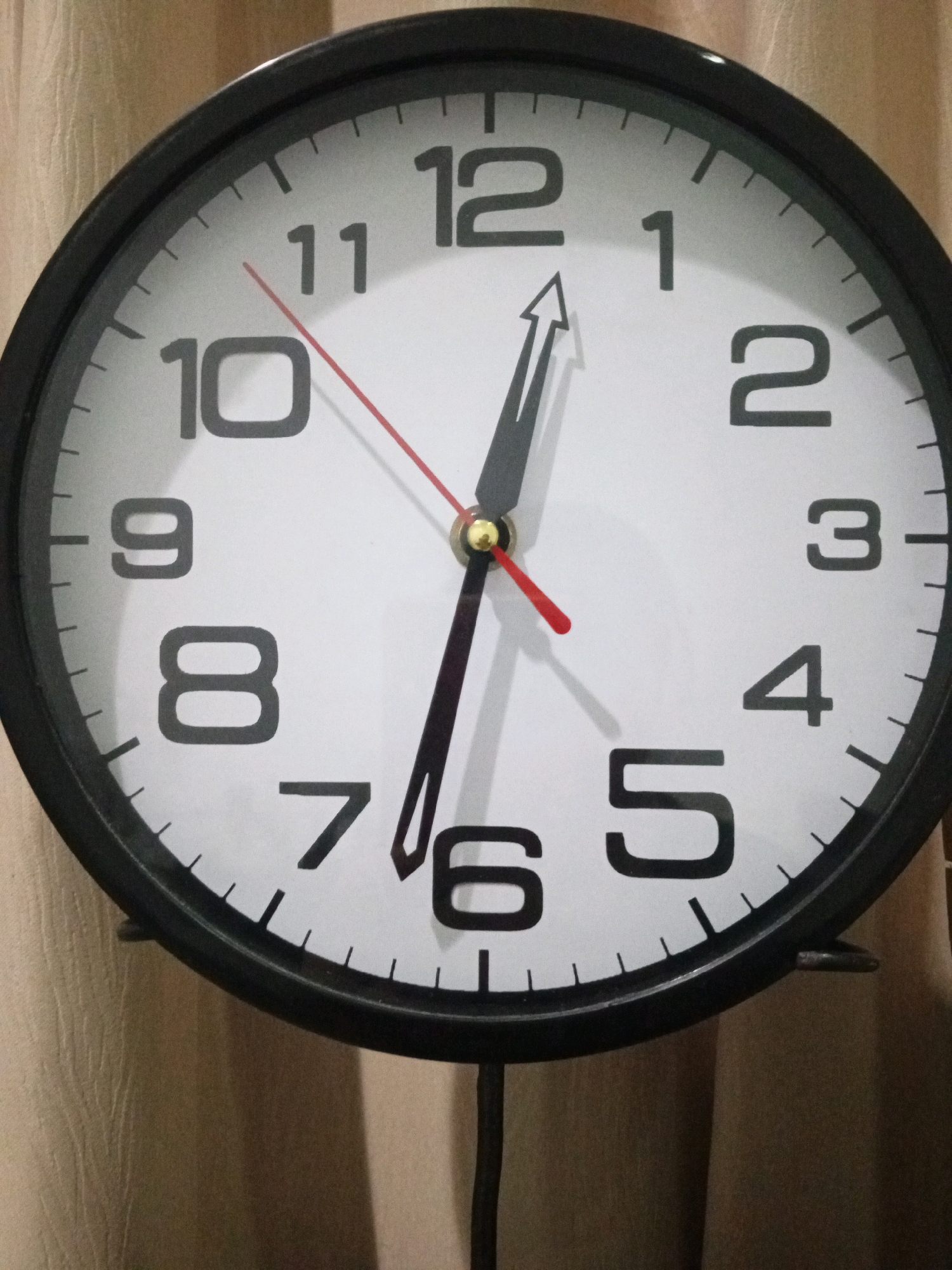 A close-up, slightly angled shot shows a round analog clock with a black frame against a beige, textured background. The clock face is white with black Arabic numerals marking the hours from 1 to 12. The clock has three hands: a black hour hand, a black minute hand, and a thin red second hand. The hour hand is pointing just past the 12, and the minute hand is pointing to the 1. The second hand is pointing to the 7. The image does not provide any information about the location in Denpasar, Indonesia.
