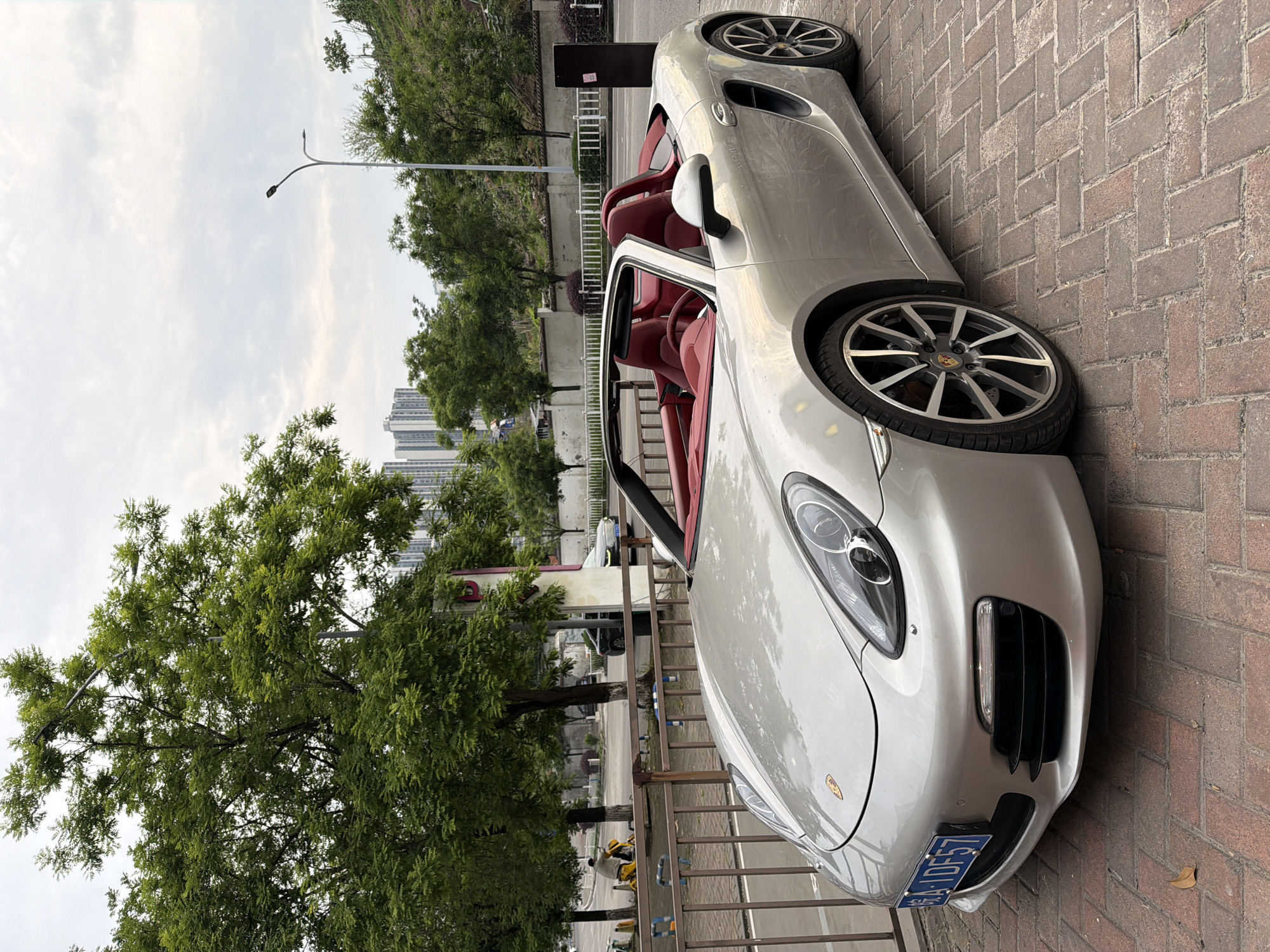 A silver Porsche Boxster convertible with a red interior is parked on a brick walkway beside a metal fence. Behind the fence are trees, a street, and buildings. The car has a blue license plate with the characters "渝ADF557." The sky is cloudy. The location is likely in Chongqing High-tech Zone, China.