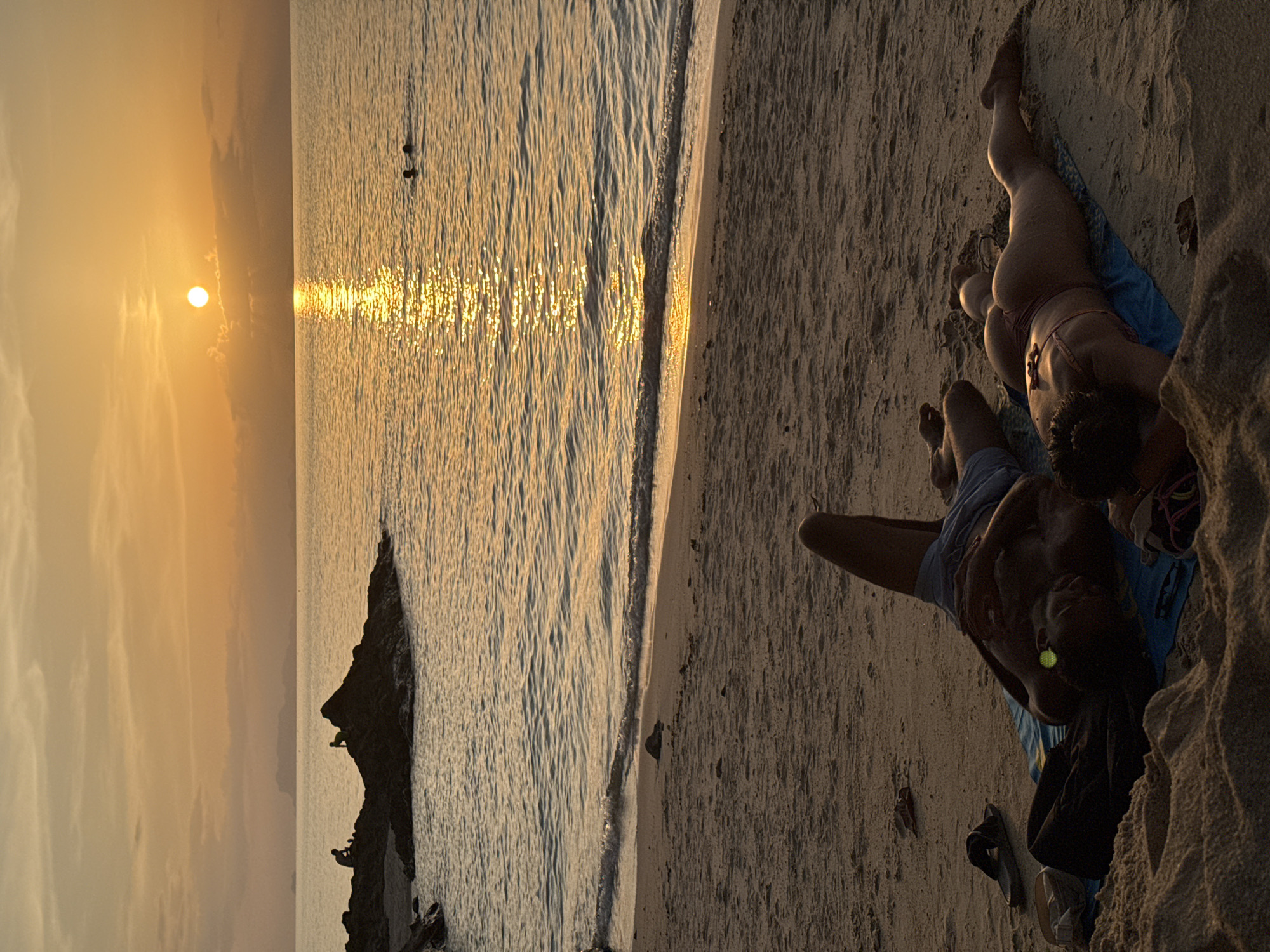 The image depicts a tranquil beach scene at sunset in Ko Samui District, Thailand. The golden hour casts a warm glow over the water, with the sun visible as a bright orb on the horizon. The sea is calm, with gentle waves lapping the sandy shore. In the foreground, two individuals are seen relaxing on towels on the beach. One person, lying on their back, appears to be a male wearing blue shorts, with a towel draped over their legs. The other person, lying face down, appears to be a female wearing a bikini. Further out in the water, two other figures are visible, suggesting they are swimming. In the distance, rocky outcrops emerge from the sea, with a few silhouetted figures perched on them. The overall atmosphere is peaceful and serene, characteristic of a tropical beach at dusk.