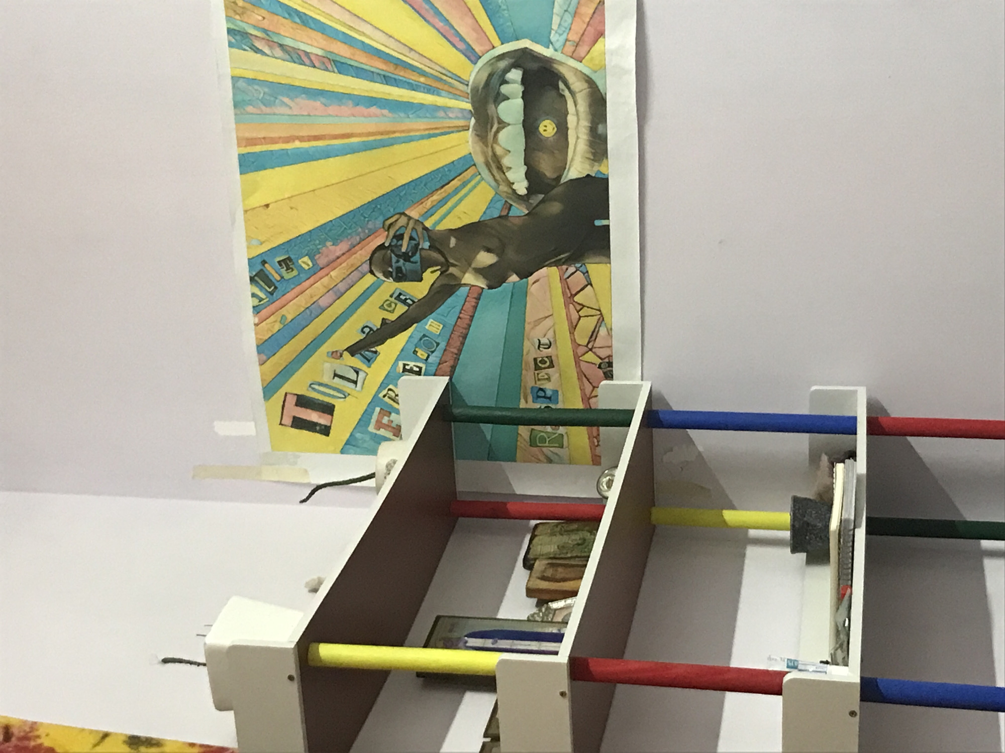 A close-up shot shows a white, three-tiered shelf with colored rods (red, yellow, green, blue) acting as supports. The shelf contains miscellaneous items, including what appears to be currency. Behind the shelf hangs a poster featuring an illustrated figure with a large open mouth above them. The walls behind the shelf are light-colored. Based on similar images from the region, the image is likely located in Nha Trang, Vietnam.