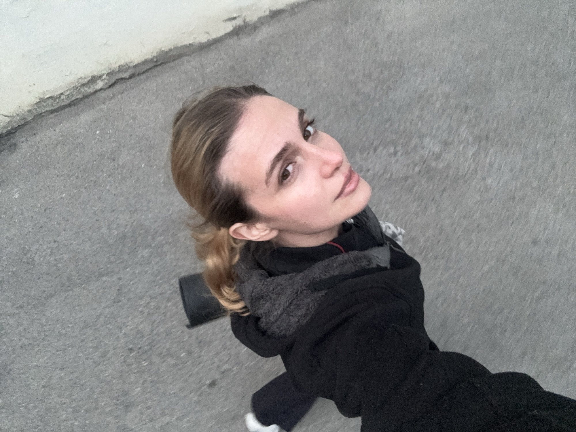 A young woman is depicted from an overhead perspective, looking upwards towards the camera, suggesting a selfie or a shot taken from above. She has light brown hair pulled back from her face, with defined eyebrows and warm brown eyes, exhibiting a calm and slightly observant expression. She is dressed in a black jacket featuring a grey faux fur or fleece lining around the collar. A dark, rectangular object, possibly a bag or part of her attire, is visible behind her left shoulder, and a glimpse of white footwear can be seen near the bottom of the frame.
The setting appears to be outdoors, likely a sidewalk or street in Tbilisi, Georgia, given the grey, textured ground. A light-colored, weathered wall or curb runs along the top left edge of the image. The lighting is soft and even, indicating either an overcast sky or an early morning/late afternoon time of day. The woman seems to be either standing still or walking, captured in a moment of upward gaze. There is no visible text in the image.