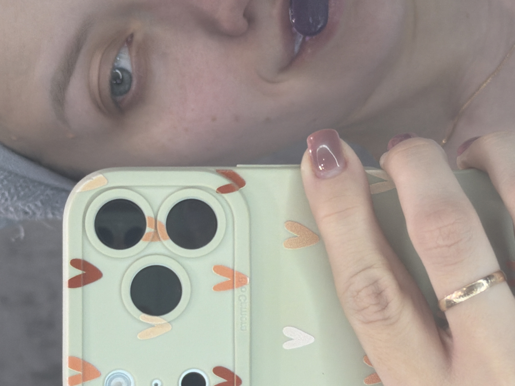 A close-up, tilted shot features a fair-skinned person holding a light green iPhone with a patterned case. The case has heart shapes and circular camera lens cutouts. The person's hand has a gold ring on the ring finger, and their nails are manicured with a mauve color. Part of the person's face is visible in the background, including an eye and a partially open mouth with a dark object inside. A gold chain is visible around the person's neck. The location of the image is unknown and cannot be confirmed to be Reshetnikovo, Russia.