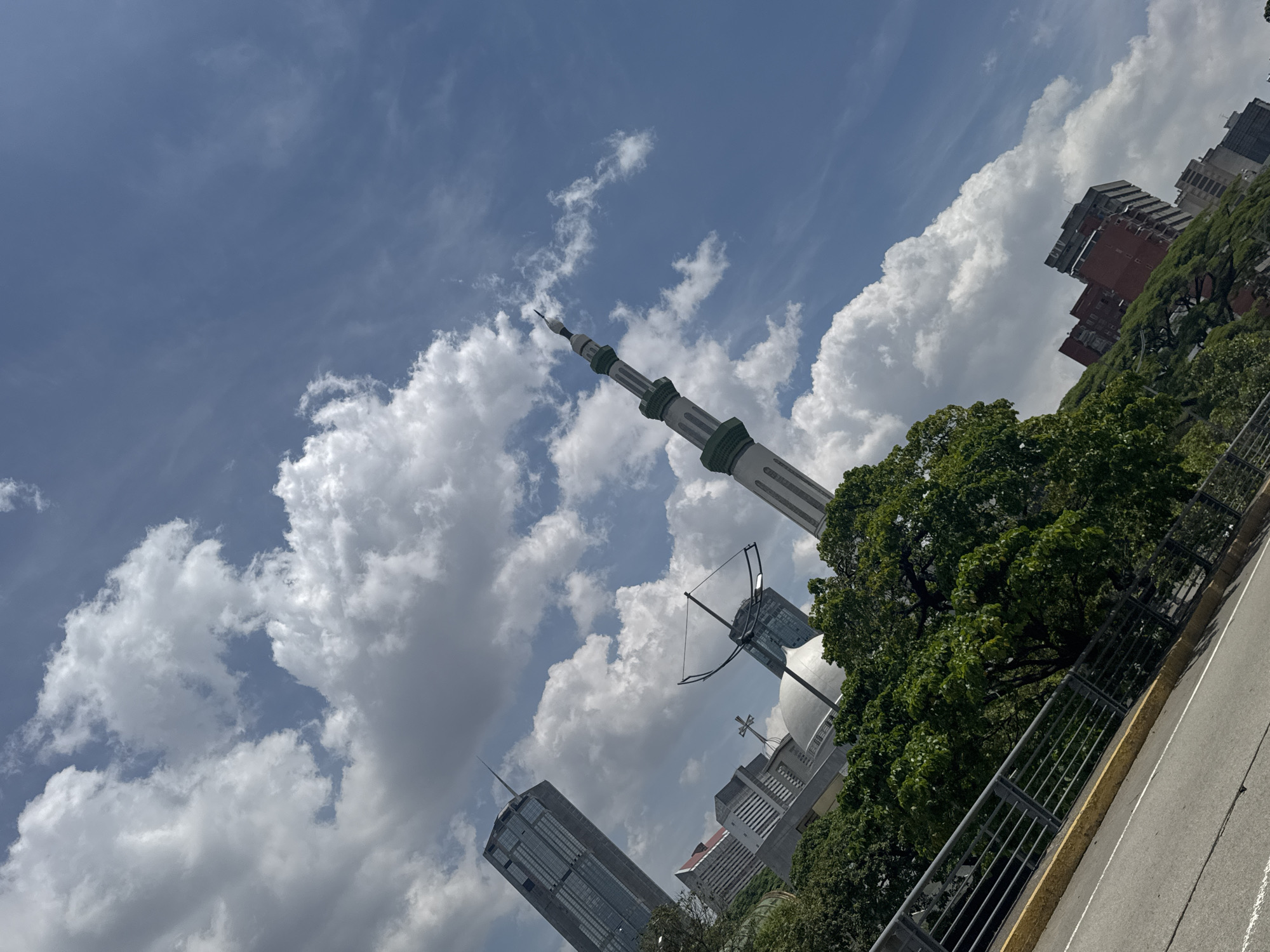 The image presents an upward, angled view of Caracas, Venezuela. A tall, cylindrical minaret with alternating bands of light and dark coloration stands prominently against a backdrop of cumulus clouds and blue sky. Adjacent to the minaret, partly obscured by trees, is a mosque with a dome and cross, signifying the coexistence of religions. Modern skyscrapers are visible in the background, interspersed with dense green foliage. A section of a road with a metal railing is in the foreground, further emphasizing the urban environment. The buildings are densely packed, indicating a high-density urban area.