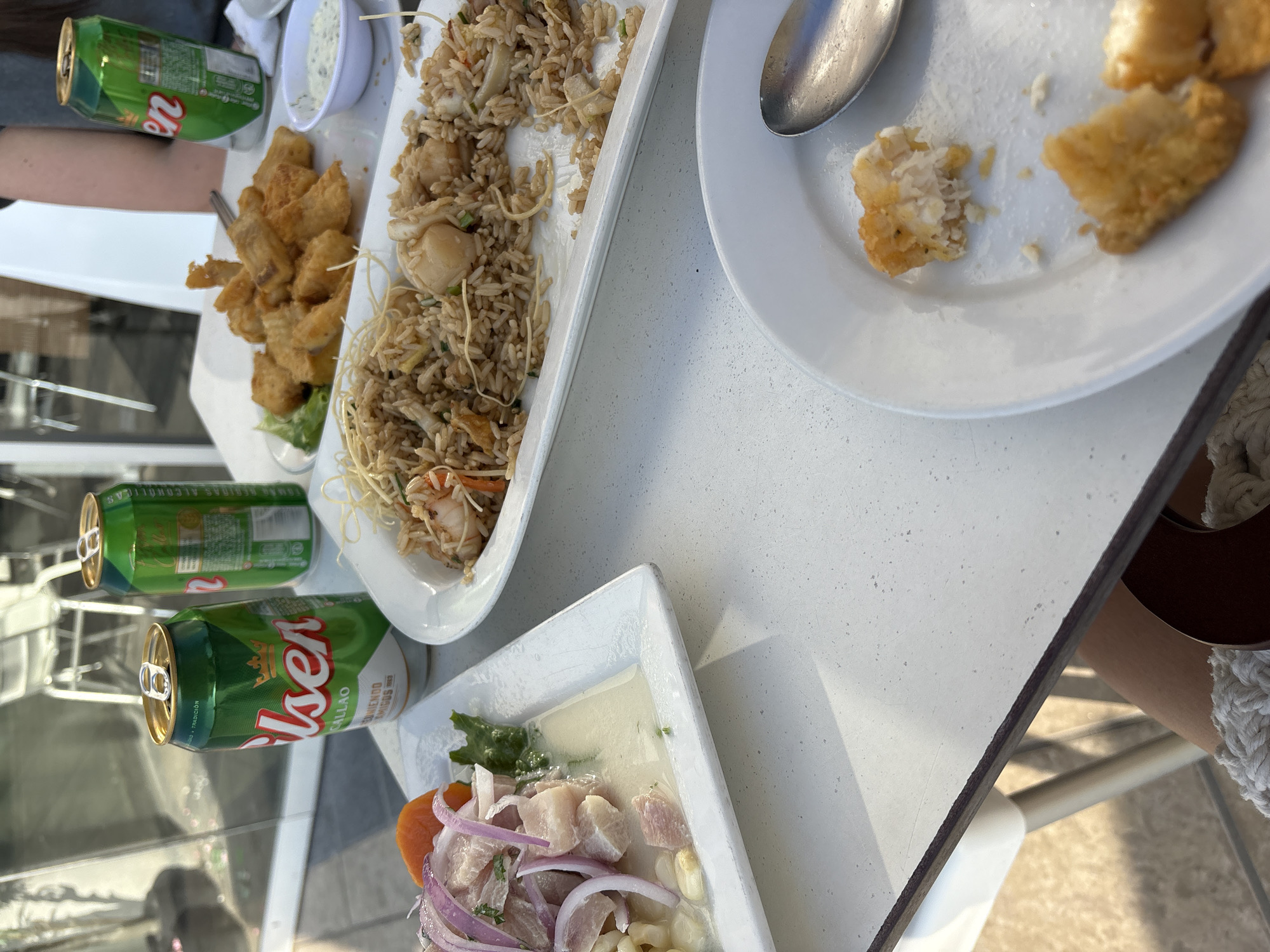 The image depicts a close-up, eye-level shot of a meal laid out on a white speckled outdoor table. Two green cans of Pilsen Callao beer are visible on the left, one opened. To the right of the beer cans, a rectangular white plate contains a portion of fried fish. Adjacent to this is a long white plate filled with fried rice, garnished with crispy noodles and containing pieces of seafood such as shrimp and scallops. In the foreground, a round white plate holds a small portion of fried fish, partially eaten, with a silver spoon resting beside it. Another white plate to the left holds a dish of ceviche, featuring chunks of white fish, red onion slivers, corn kernels, and a slice of orange. The setting appears to be an outdoor dining area, possibly a restaurant terrace, with hints of blurred outdoor seating and natural light suggesting a daytime meal. The overall scene conveys a casual dining experience.