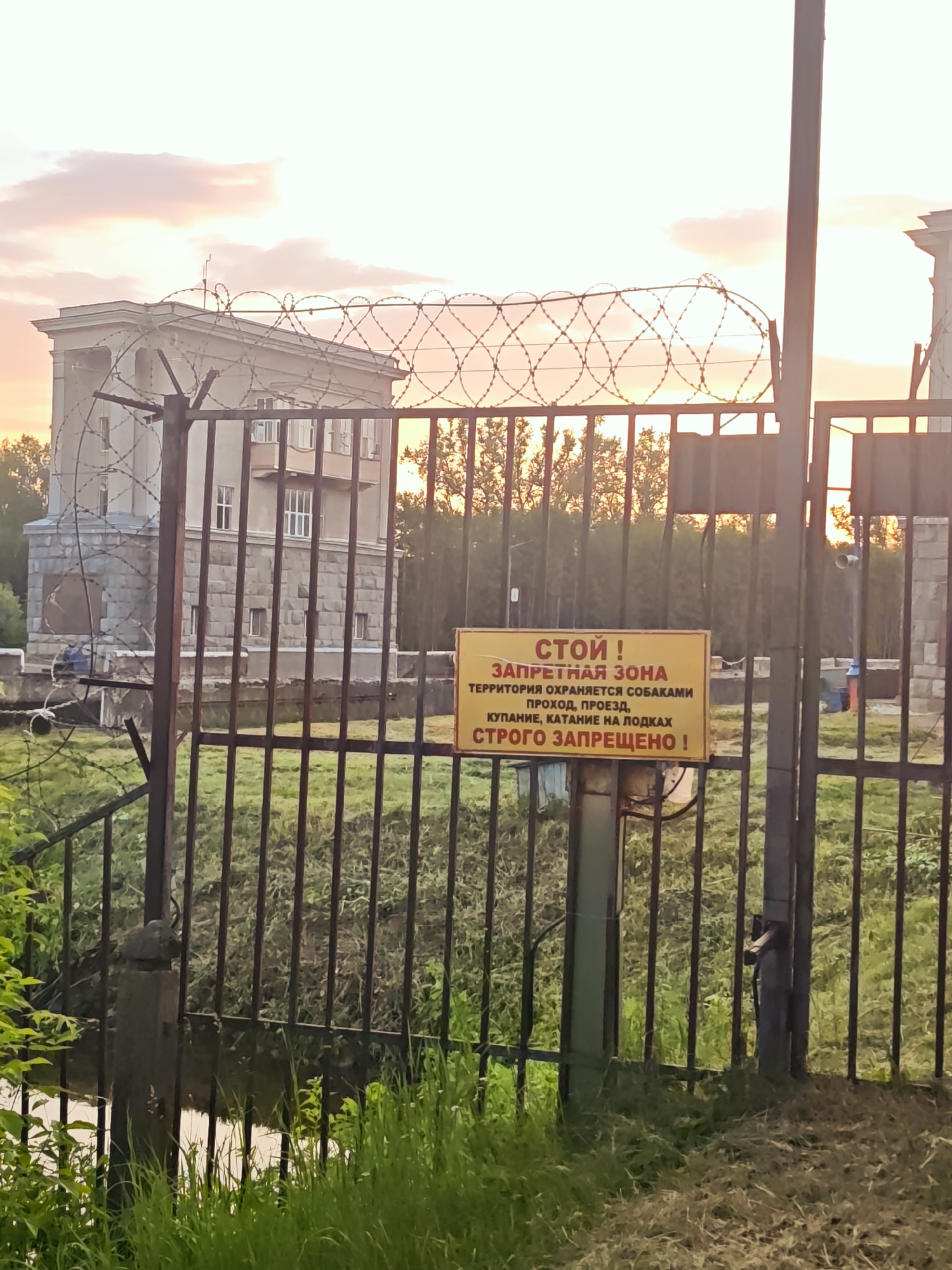 A rusty metal gate with barbed wire along the top is in the foreground. Behind the gate, a yellow sign with red and black lettering is attached to a metal pole. The sign reads: "СТОЙ! ЗАПРЕТНАЯ ЗОНА ТЕРРИТОРИЯ ОХРАНЯЕТСЯ СОБАКАМИ ПРОХОД, ПРОЕЗД, КУПАНИЕ, КАТАНИЕ НА ПОДКАХ СТРОГО ЗАПРЕЩЕНО!" This translates to: "STOP! PROHIBITED ZONE THE TERRITORY IS PROTECTED BY DOGS PASSAGE, TRAVEL, SWIMMING, RIDING ON TUBES IS STRICTLY PROHIBITED!"
Behind the gate and sign, a large, imposing concrete building with multiple windows is visible. The sky above is filled with soft, pastel-colored clouds, suggesting either sunrise or sunset. Trees and green grass surround the building and the gate. The overall scene appears to be in an industrial or fenced-off area within the city of Vodnik Snt, Russia.