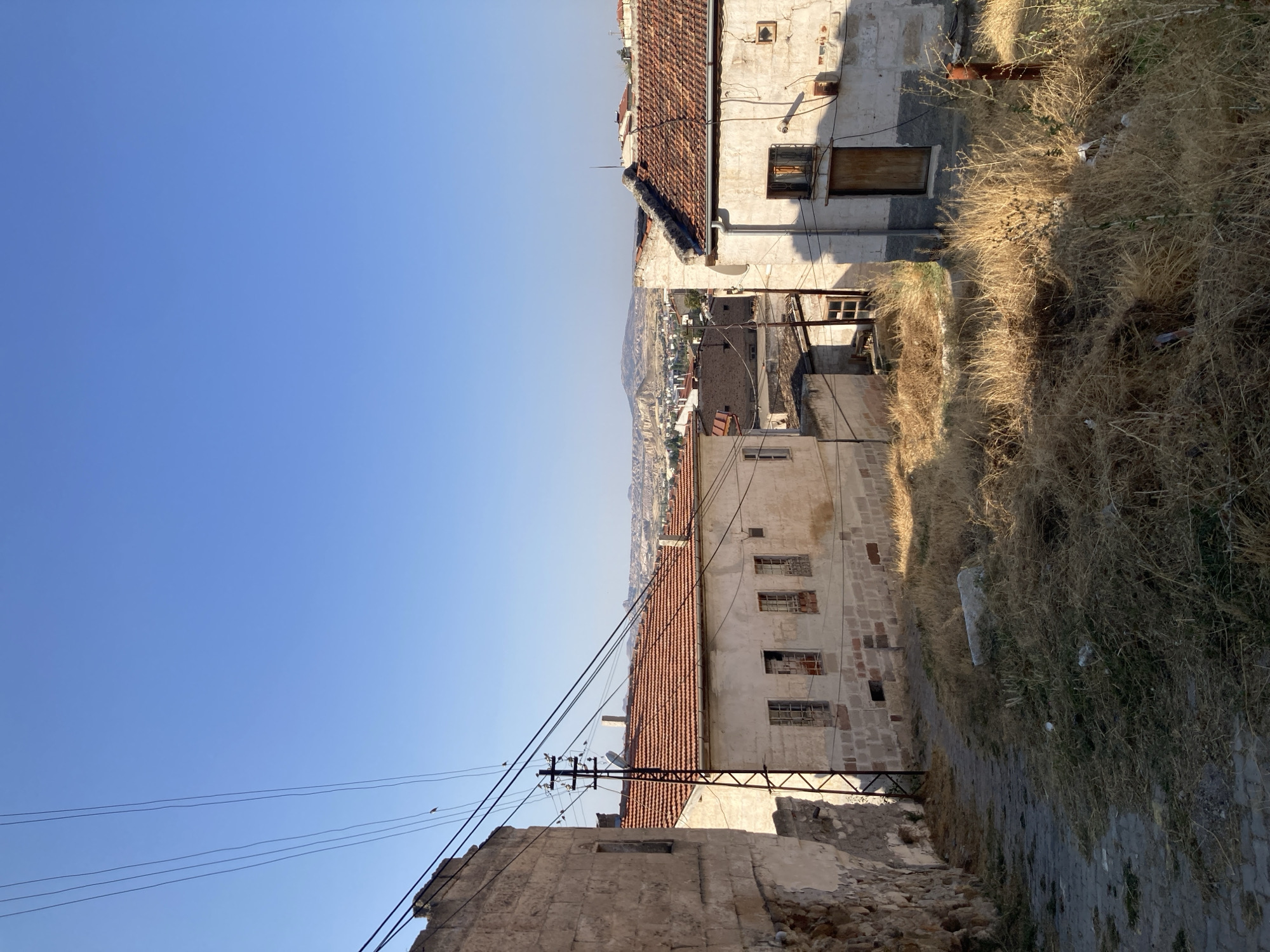 The image depicts a view looking down a narrow, cobblestone street in Avanos İlçe Merkezi, Turkey. The street is lined with old, weathered buildings constructed from stone and plaster, many of which appear to be abandoned or in a state of disrepair. The architecture suggests traditional Anatolian style, characterized by red-tiled roofs and simple facades.
On the left side of the street, a stone wall of an older building is visible, with a section of its exterior crumbling away. Further down the street, a building with multiple windows, some boarded up and others broken, is prominently featured. The windows are framed with metal grates. Across the street, another building stands with a wooden door and a single, small window. The plaster on this building is cracked and stained, and a patch of what appears to be a framed picture hangs crookedly on the exterior wall.
The foreground and sides of the street are overgrown with dry, yellowed grass and weeds, suggesting a lack of recent maintenance. In the distance, the cityscape of Avanos can be seen, with more buildings and the characteristic geological formations of Cappadocia. A clear blue sky stretches across the upper portion of the image, with a few power lines strung across the view, supported by a utility pole. The lighting indicates it is daytime, likely late morning or early afternoon, given the strong shadows cast by the buildings. There are no people visible in the scene. The overall impression is one of a quiet, historical street in a town known for its unique landscape and traditional architecture.