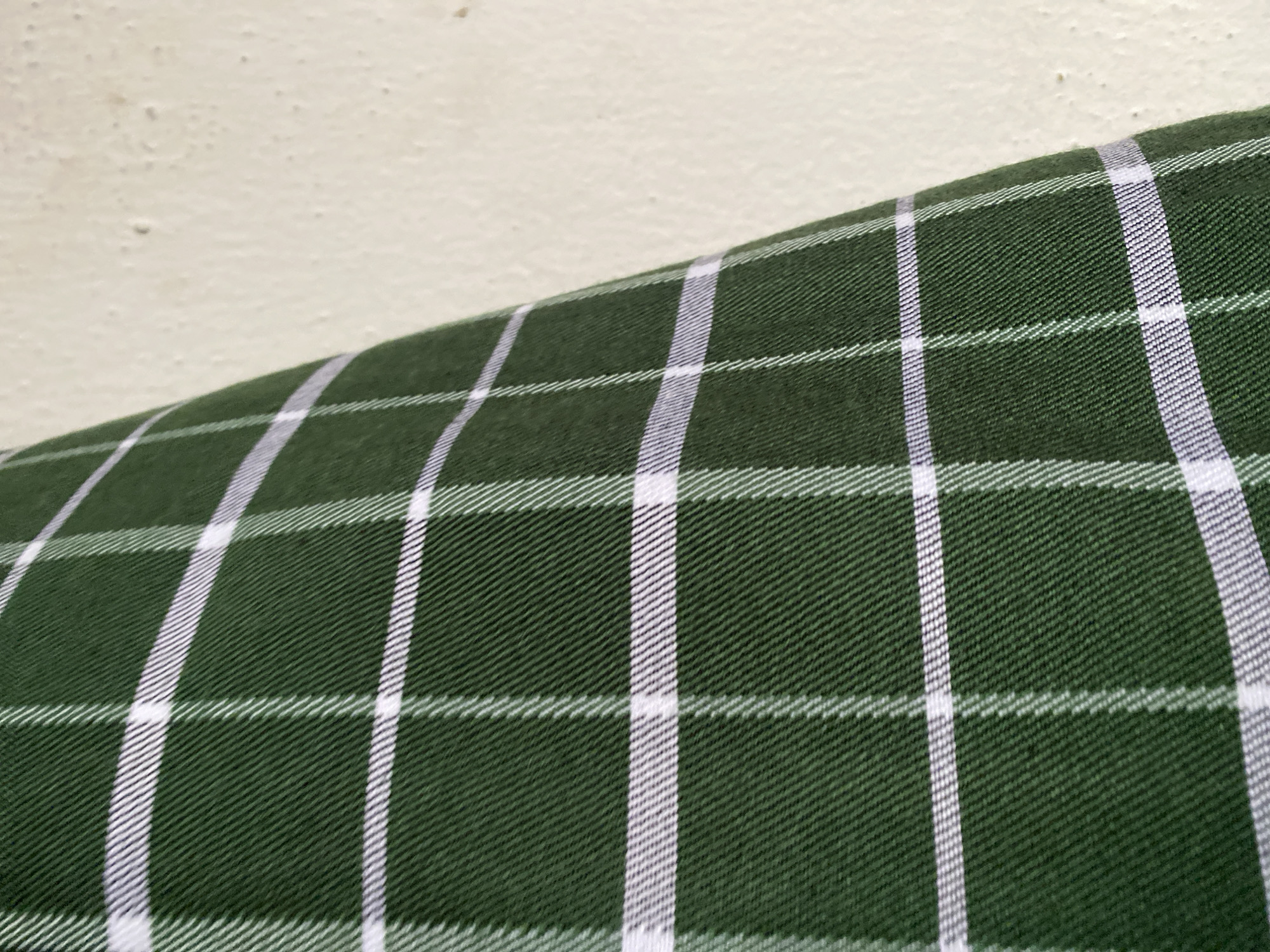 The image shows a close-up of a green plaid fabric with thin white lines forming a checkered pattern. The fabric appears to be a soft material, possibly cotton or flannel. It is draped over a textured off-white surface, which could be a wall. The lighting is even, highlighting the texture of the fabric and the surface. There are no people or specific objects visible beyond the fabric and the background surface. The image is abstract and focuses on the pattern and texture. No specific location within Jauro Yinu, Nigeria can be determined from the visual information provided.