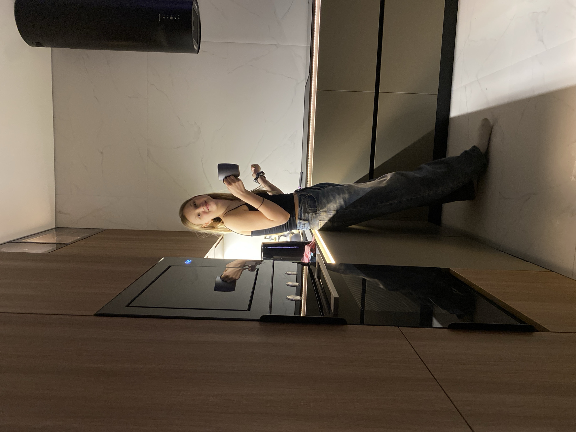 A young woman stands in a modern kitchen in what is likely Chaikovsky, Russia, based on the query. She holds a dark-colored mug in her hands. She wears a black tank top and blue jeans, her blonde hair loosely framing her face. The kitchen features light-colored marble-like backsplash, dark, possibly wood-look cabinets and drawers, and stainless steel appliances, including an oven with a stovetop above. The lighting is a mix of natural and artificial. The oven's surface reflects the overhead lighting and the woman's form. A black cylindrical vent hood hangs above the stovetop. The woman appears relaxed and casual in the kitchen setting.
