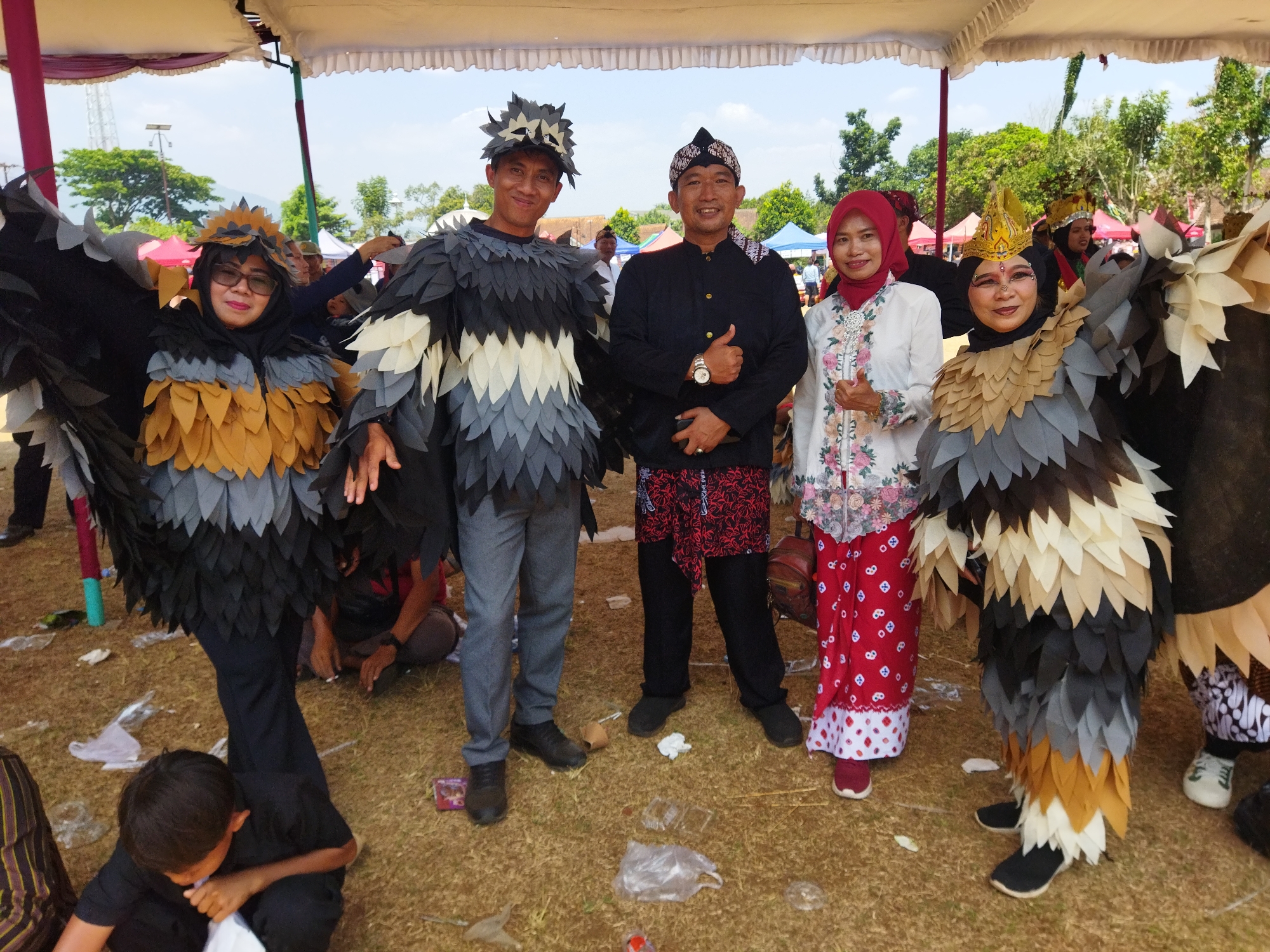 The image shows a group of people in costumes resembling birds, possibly as part of a cultural event or parade in Semarang, Indonesia. The main subjects are four individuals standing close together, two on the left and two on the right, with a fifth person visible on the far right. The two individuals on the left are dressed in elaborate bird-like costumes with layered, feather-shaped elements in black, gray, and tan colors. One is wearing a dark headscarf and glasses, and the other wears a spiky headpiece resembling a bird's crest. The two individuals on the right are also in elaborate bird costumes. One wears a red hijab and a white embroidered top with a patterned red skirt. The person next to them, on the far right, is also in a bird costume. Two men in traditional Indonesian attire, a black long-sleeved shirt and pants with a patterned sarong, are standing between the bird-costumed individuals. The ground is covered in dry grass and scattered litter, suggesting an outdoor event. In the background, there are tents and trees, indicating a setting in a park or open field. A child in a black shirt is partially visible in the lower left foreground, looking down at something in their lap. The overall scene appears to be a festive or ceremonial gathering.