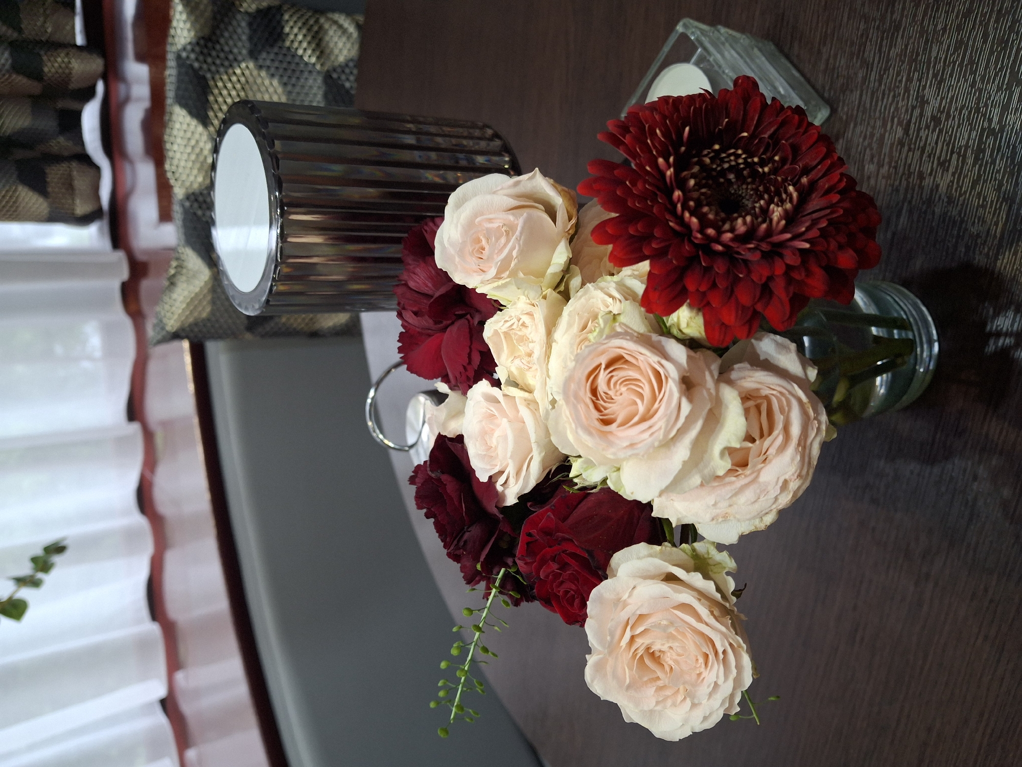 A vase of flowers, including blush roses, dark red roses, and a red gerbera daisy, sits on a dark wooden table. A decorative glass cylinder with vertical ridges is positioned behind the flowers. To the right of the flowers, a small clear glass cube holding a white candle is visible. The scene is set indoors, likely in a restaurant or cafe, given the presence of upholstered seating and curtains in the background, suggesting an urban setting. This setting could be Pila, Poland.