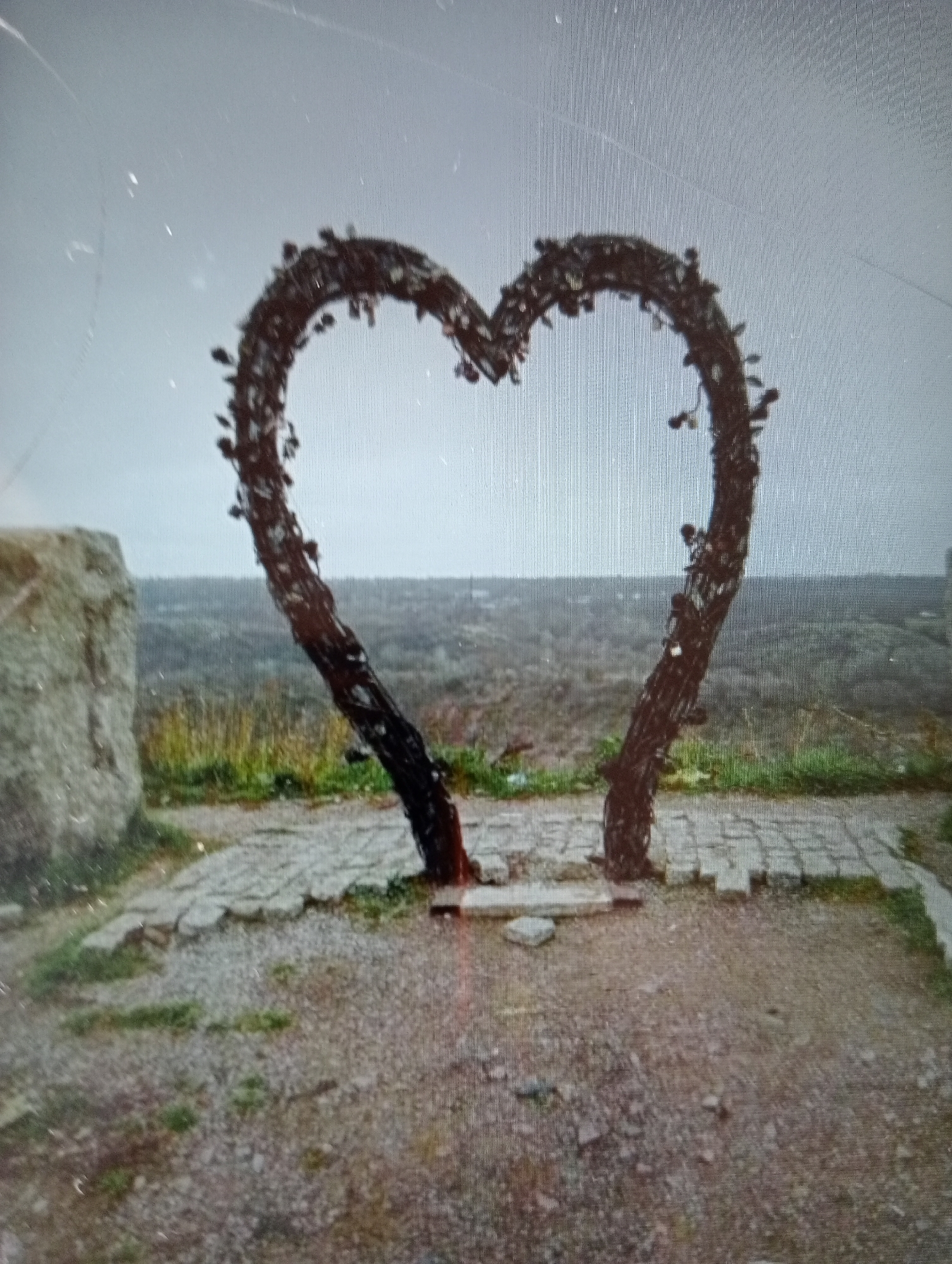 A heart-shaped sculpture made of dark, twisted branches stands in the foreground of an outdoor scene. The sculpture is adorned with small, dark leaf-like elements. To the left of the sculpture is a large, rough-hewn stone. The ground in front of the sculpture is paved with irregular flagstones and gravel, with patches of sparse grass. In the background, a hazy landscape of rolling hills and vegetation stretches to the horizon under a pale, overcast sky. The scene appears to be in a natural or park-like setting.