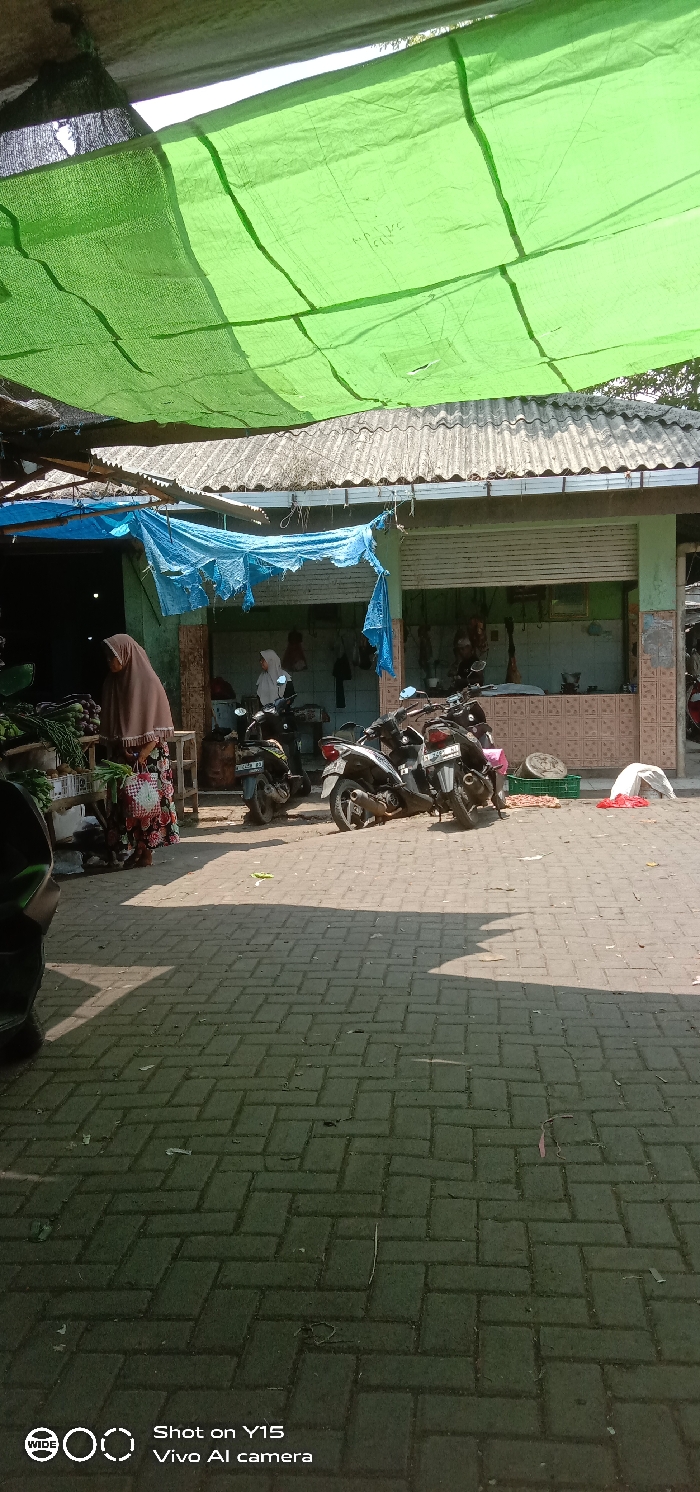 The image displays an outdoor market scene in Maron, Indonesia. Overhead, a large, light-green tarpaulin provides shade. Beneath this, a building with a light blue-green facade and a corrugated metal roof is visible, with a dilapidated blue tarp draped below its roofline.
In the left foreground, a woman wearing a brown hijab and a patterned skirt stands beside a market stall displaying various fresh vegetables, including eggplants. A red and white checkered bag is positioned on the ground near her feet.
The building's entrance, to the right of the stall, features a partially open metal roll-down shutter above a tiled half-wall with a brown geometric pattern. Inside this opening, two individuals are present: one woman in a light-colored hijab stands near a counter, and another figure is further inside, positioned near several suspended meat carcasses.
Four motorcycles are parked on the interlocked brick pavement in the mid-ground. These vehicles include dark-colored, silver, and black models. Visible license plates include "B 2450 NJ" and "B 6458 DJ." A fifth motorcycle is partially visible in the extreme left foreground.
On the ground near the building, a green plastic crate, a circular metal basin, a white bag, and a red plastic bag are present. The paved surface shows scattered small debris. Defined shadows are cast across the pavement, indicating direct sunlight.