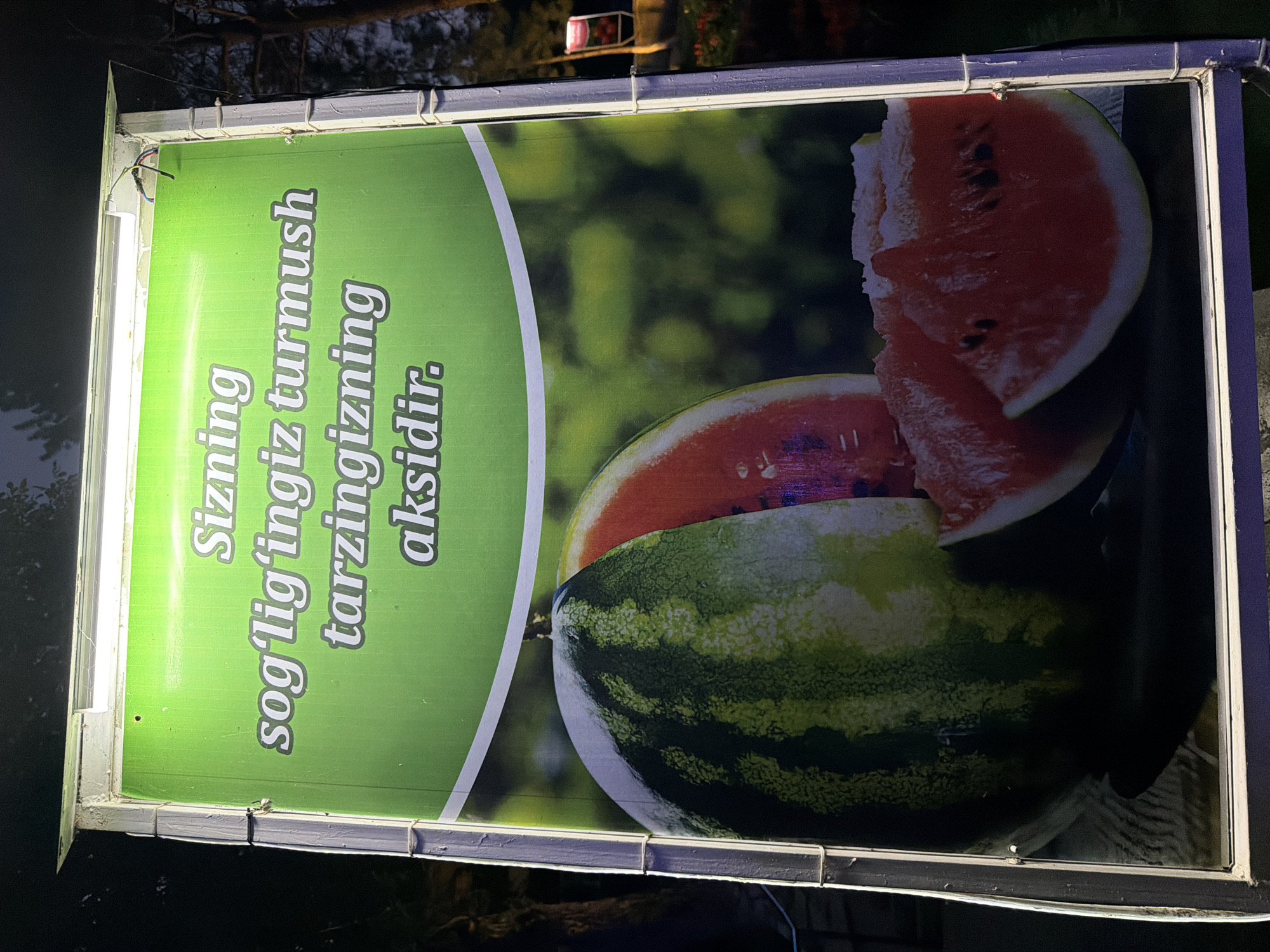 A brightly lit sign in Khumsan, Uzbekistan, features a vibrant green background with white text and an image of watermelons. The text, written in Uzbek, translates to "Your health is a reflection of your lifestyle." The top portion of the sign has a white border with lighting elements visible. Below the text, a large, whole watermelon is prominently displayed on the left, with its rind showing dark green stripes. To the right of the whole watermelon, a cut watermelon is visible, revealing its red flesh and black seeds. Two slices of watermelon are also arranged in the foreground, also showing their red interior and seeds. The background of the sign is a soft-focus green, suggestive of foliage. The sign is illuminated from above by fluorescent lights, casting a glow on the scene. The sign is framed by a light-colored metal structure, with visible wiring and mounting hardware. The environment surrounding the sign is dark, indicating it is nighttime. In the background, faint lights and possibly more foliage are discernible.