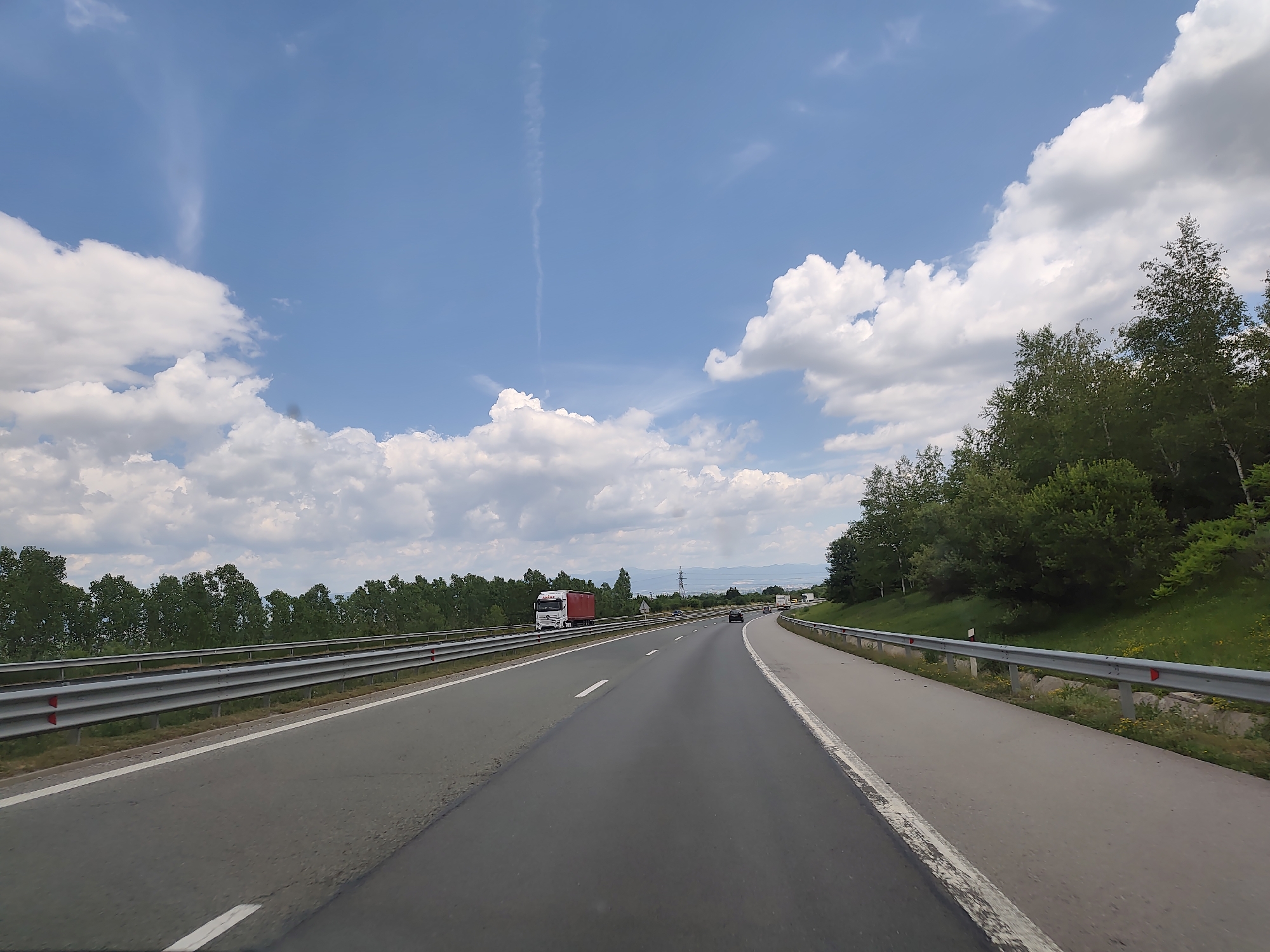The image depicts a view from a vehicle on a highway, with the sky a vibrant blue dotted with fluffy white clouds. On the left side of the highway, a silver guardrail runs parallel to the road, with a dense line of green trees beyond it. A white truck with a red trailer is visible on the highway ahead. The right side of the highway is bordered by a grassy embankment covered in trees. In the distance, mountains are visible under the hazy sky. The road itself is paved asphalt with white lane markings. The overall scene suggests a sunny day on a rural highway. The location is confirmed as Malo Buchino, Bulgaria, by the context provided.