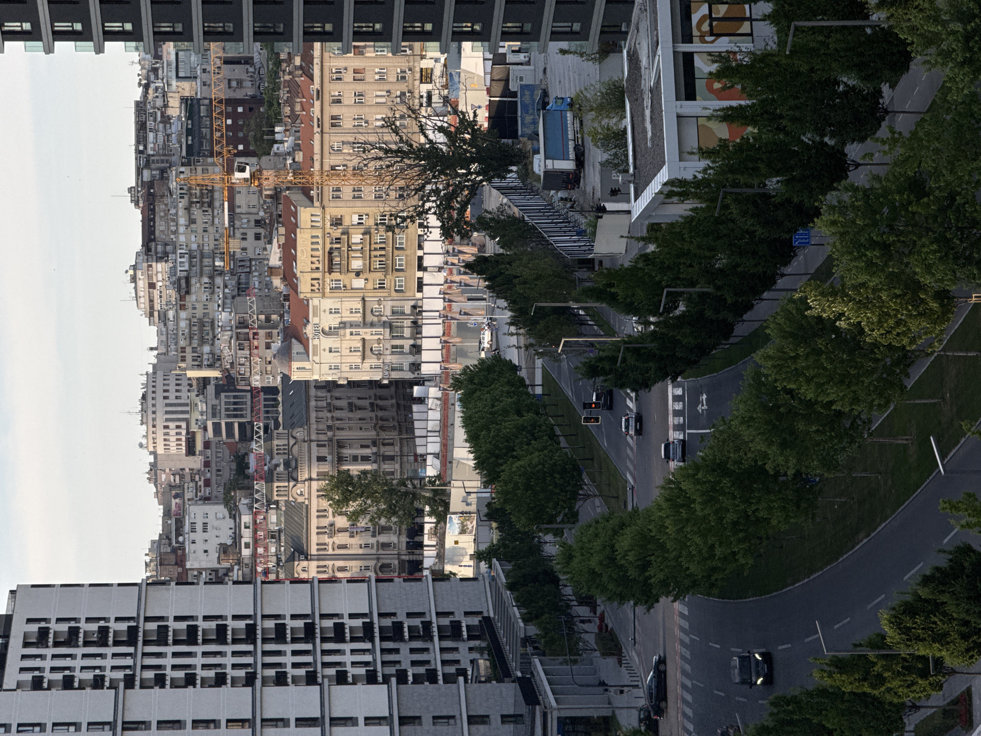 The image shows an elevated view of a city with a mix of old and modern buildings. A major road lined with trees runs through the scene. Several cars are visible on the road. Construction cranes are present amidst the older buildings.