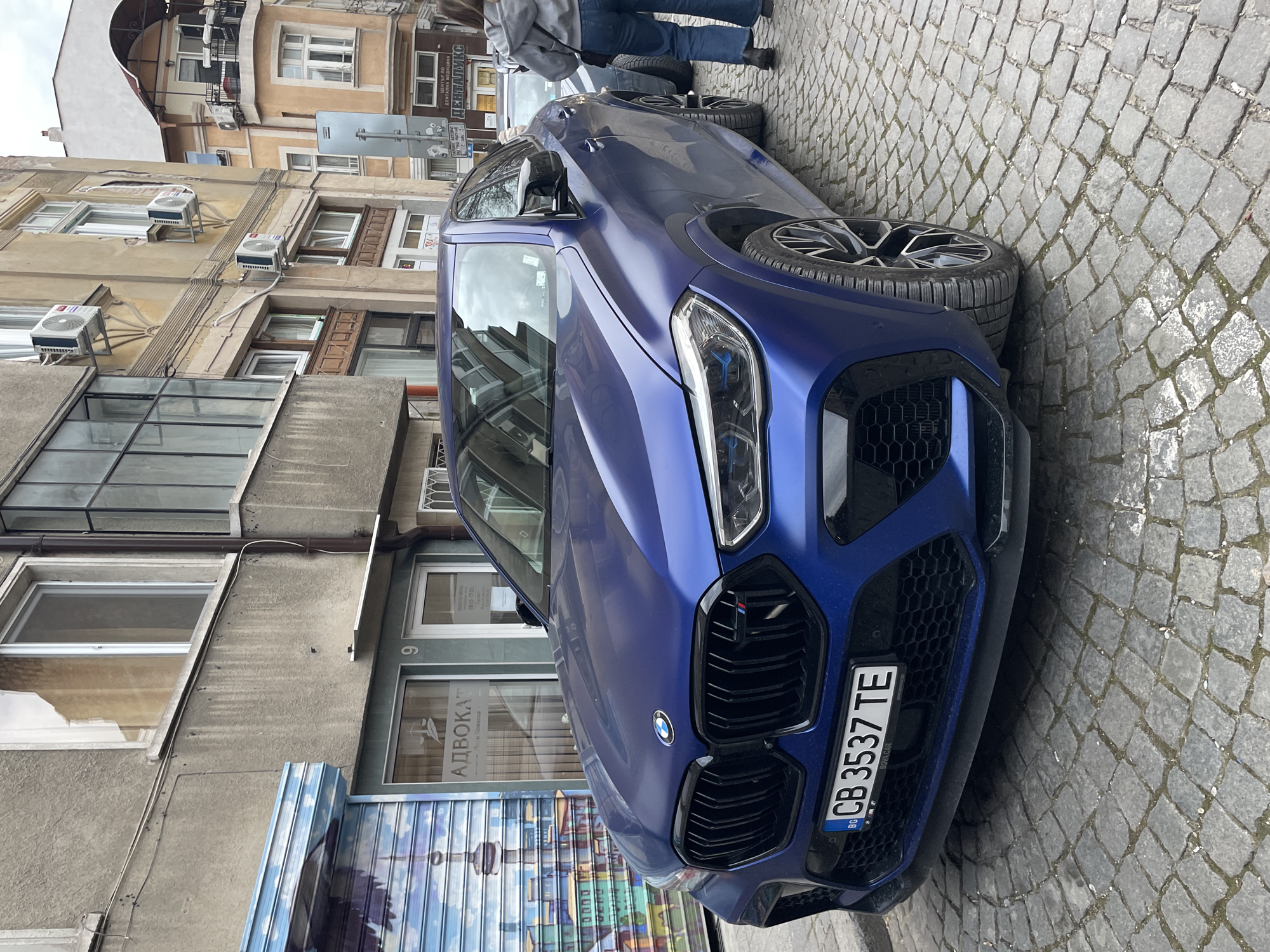 The image depicts a blue BMW X6 M Competition parked on a cobblestone street in Plovdiv, Bulgaria. The car's license plate reads "CB 3537 TE." Behind the car is a building with a sign for "АДВОКАТ" (lawyer) and the name "Анри Божкова" with working hours. The number 9 is visible next to the sign. Above, there are several air conditioning units attached to the building's facade. A person wearing a gray hoodie and jeans is partially visible near the back of the car. The street is made of cobblestones. There is another car parked near the BMW.