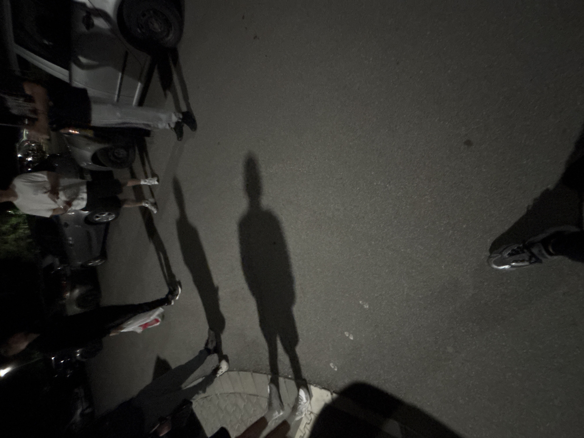 The image is a tilted, high-angle view of a dark street or pavement. The scene appears to be lit by artificial light sources, resulting in long shadows. Multiple people are present, indicated by their shadows and partially visible figures. Some individuals are near a vehicle, possibly a car, with a tire visible. Other objects are present, but difficult to discern due to the darkness and angle. The location is unspecified, but the user requested it be in Plovdiv, Bulgaria. Specific information to verify its location within Plovdiv, Bulgaria is not discernible from this image.