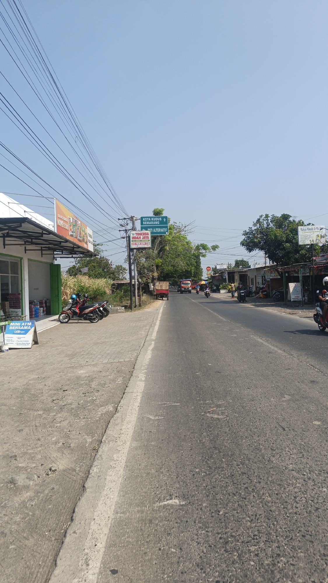 A street scene in Jekulo, Indonesia is depicted under a clear blue sky. The road, paved with asphalt and marked with a white line, stretches into the distance, with traffic including a truck, motorcycles, and other vehicles. On the left side of the road, a sidewalk is visible, lined with a building that appears to be a small store or shop, featuring awnings and signage. Several motorcycles are parked on the sidewalk in front of the building. A sign indicating "MINI ATM BERSAMA" is prominently displayed. Further down the street, more buildings and trees line the roadside, suggesting a commercial or residential area. A green directional signpost indicates "KOTA KUDUS SEMARANG" and "Dati Alternatif," suggesting this is a junction or intersection on a route leading to these cities. Power lines are strung across the upper portion of the image. The environment appears to be a moderately busy street on a sunny day.