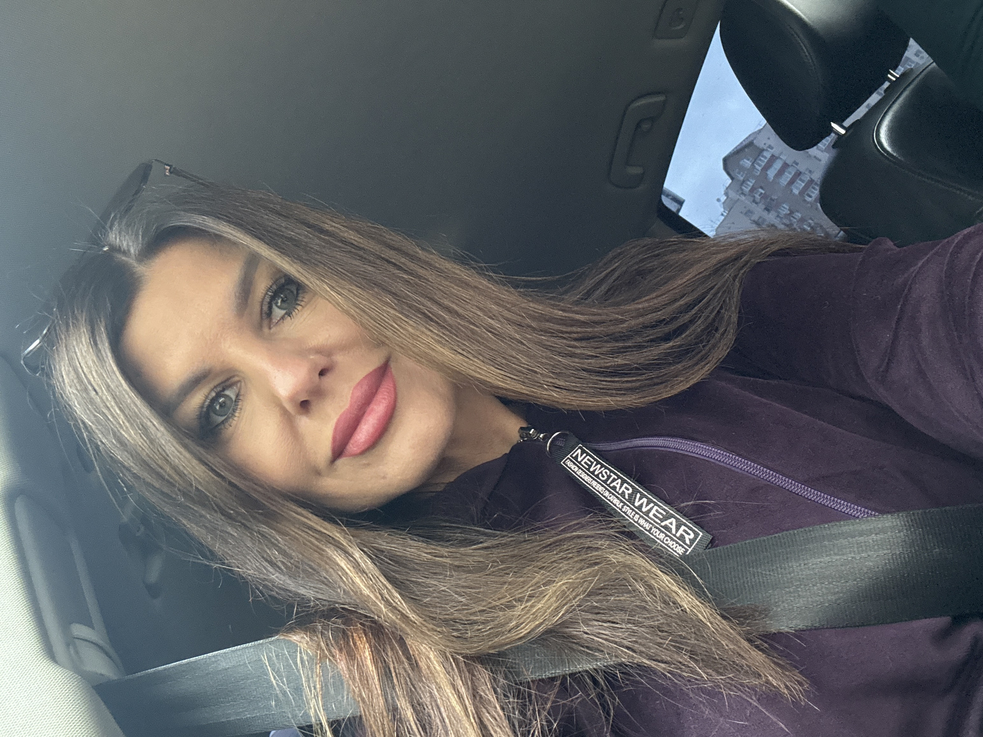 A selfie of a woman taken from a low angle inside a vehicle. She has long, brown hair that falls across her face and shoulders. She is wearing a dark purple jacket with a zipper and a tag that reads "NEWSTAR WEAR FASHION DESIGNERS PRESENT ON CATWALK STYLE IS WHAT YOUR CHOOSE". A black seatbelt is visible across her chest. Her makeup includes noticeable eyeliner and pink lipstick. The background shows the interior of a car, with the sky and a building visible through a window. The lighting suggests it might be daytime.