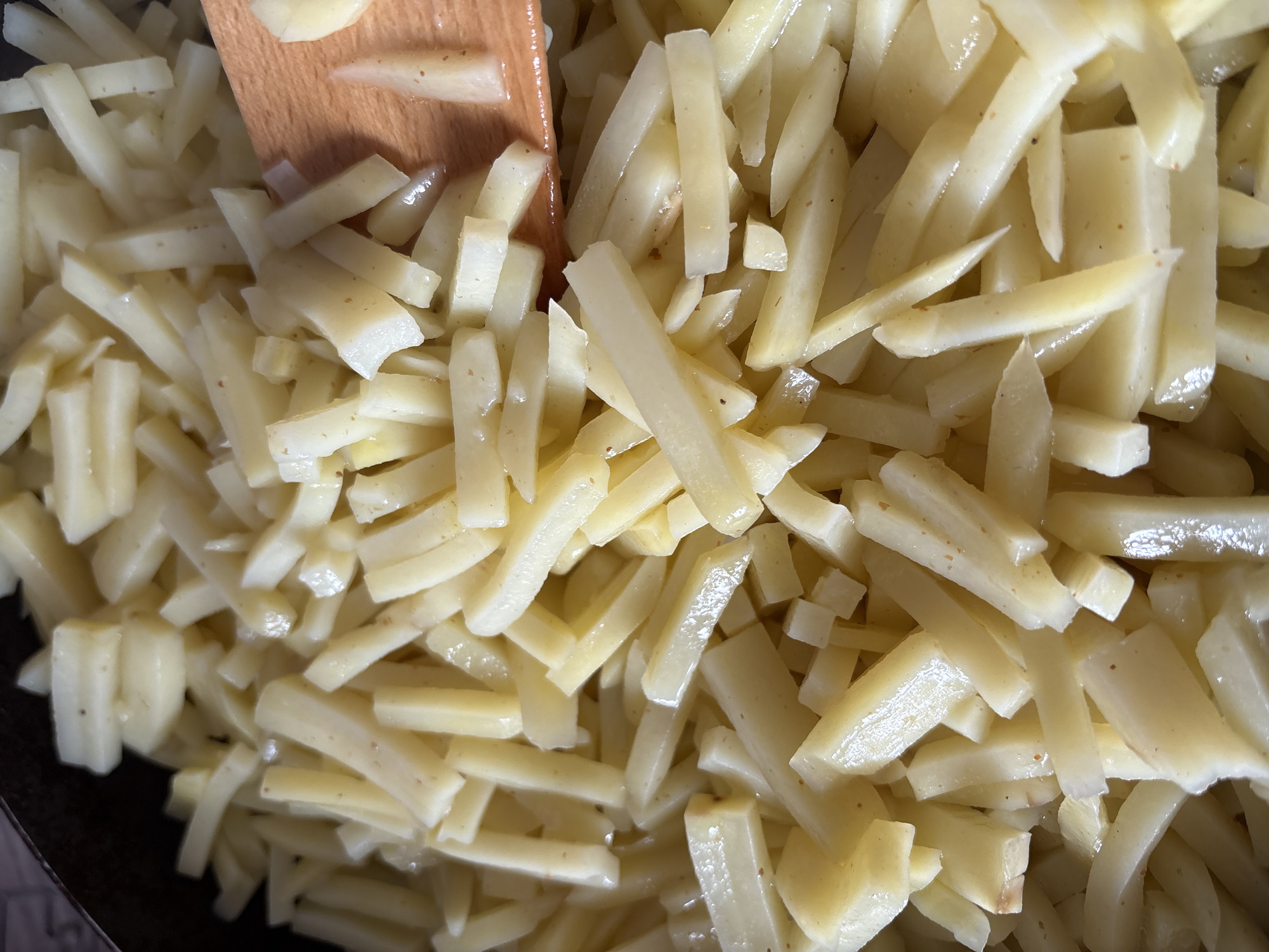 The image shows a close-up view of raw potatoes cut into french fry shapes. A wooden spatula is partially visible at the top of the frame. The potatoes are in a pan, possibly ready to be cooked. The location is not discernible from the image.