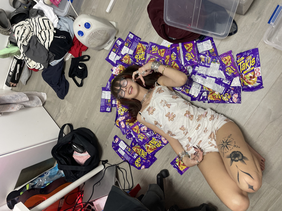 A young woman with dark hair and glasses reclines on a floor amidst numerous bags of Takis. She wears a teddy bear-patterned pajama set. Her thighs feature tattoos. The surrounding area is cluttered, featuring piles of clothes, a space heater, storage containers, and a black bag. A desk with electronic devices is also visible. The image is taken from a slightly overhead perspective, highlighting the chaotic arrangement.