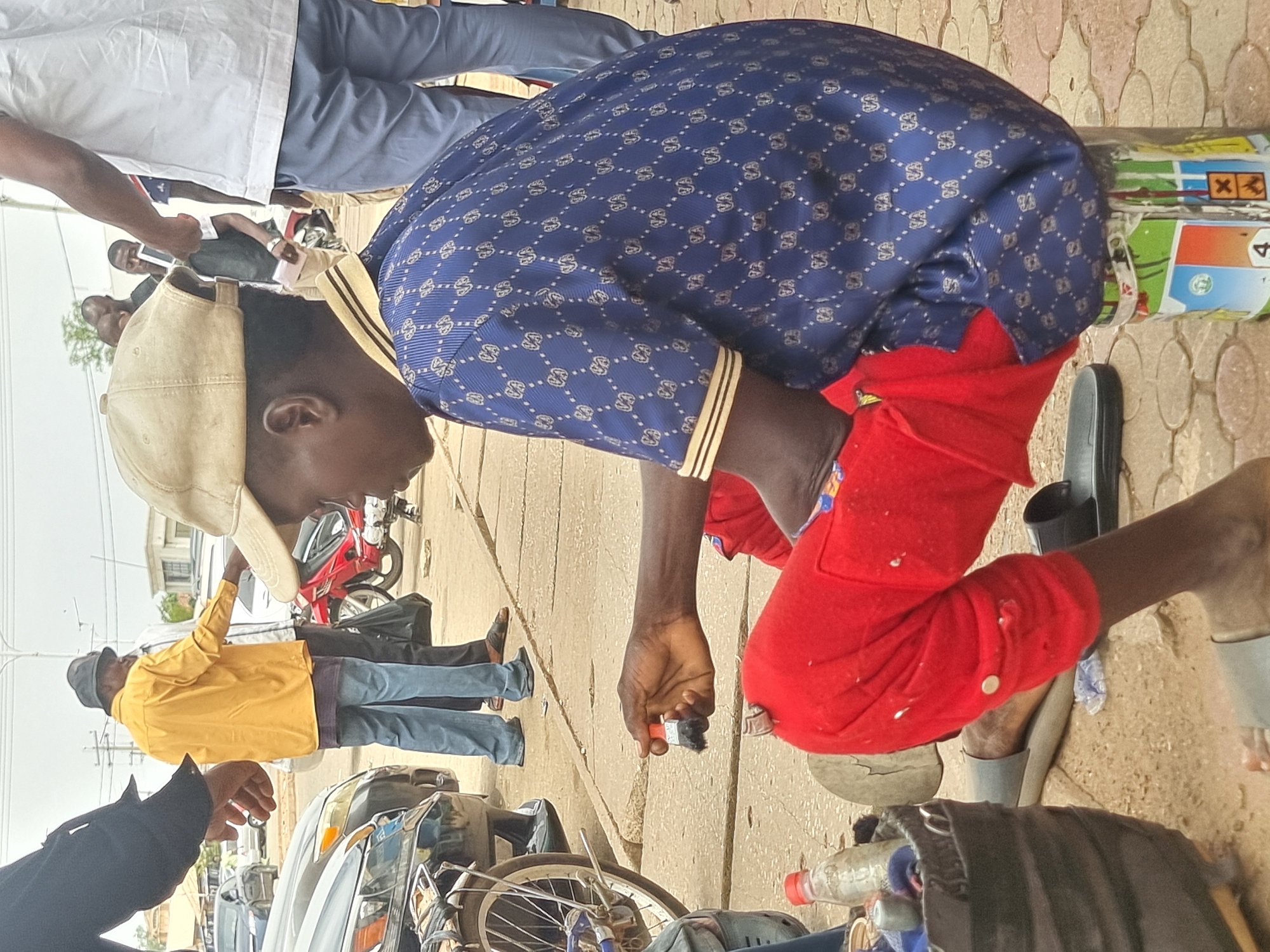In Jos, Nigeria, a man kneels on a paved surface, concentrating on an object he holds in his hand. He is wearing a blue patterned shirt, red pants, and a beige cap. Several other people are standing nearby, and a motorcycle and a car are visible in the background. The setting appears to be a busy street.
