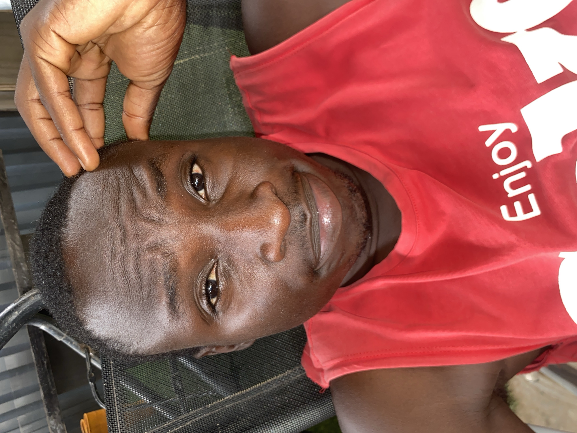 A close-up, high-angle portrait captures a man with dark skin, lying down and gazing directly at the camera with a calm, almost smiling expression. He wears a bright red sleeveless tank top that features white text, with the word "Enjoy" clearly visible and oriented vertically on the right side of his chest. His right hand is casually placed, with his fingers resting on the side of his forehead.
The man's skin exhibits a natural sheen, suggesting a warm or humid environment. His eyes are dark and alert, reflecting light. He has short, dark hair and a faint mustache and beard. He appears to be relaxing outdoors, possibly on a recliner or hammock, indicated by the black, mesh-like fabric and a partial metal frame visible beneath and to his left. The background is softly blurred, showing hints of greenery, confirming an outdoor setting during daylight hours. The overall mood is one of relaxation. The image's context is Barden Yana, Nigeria.