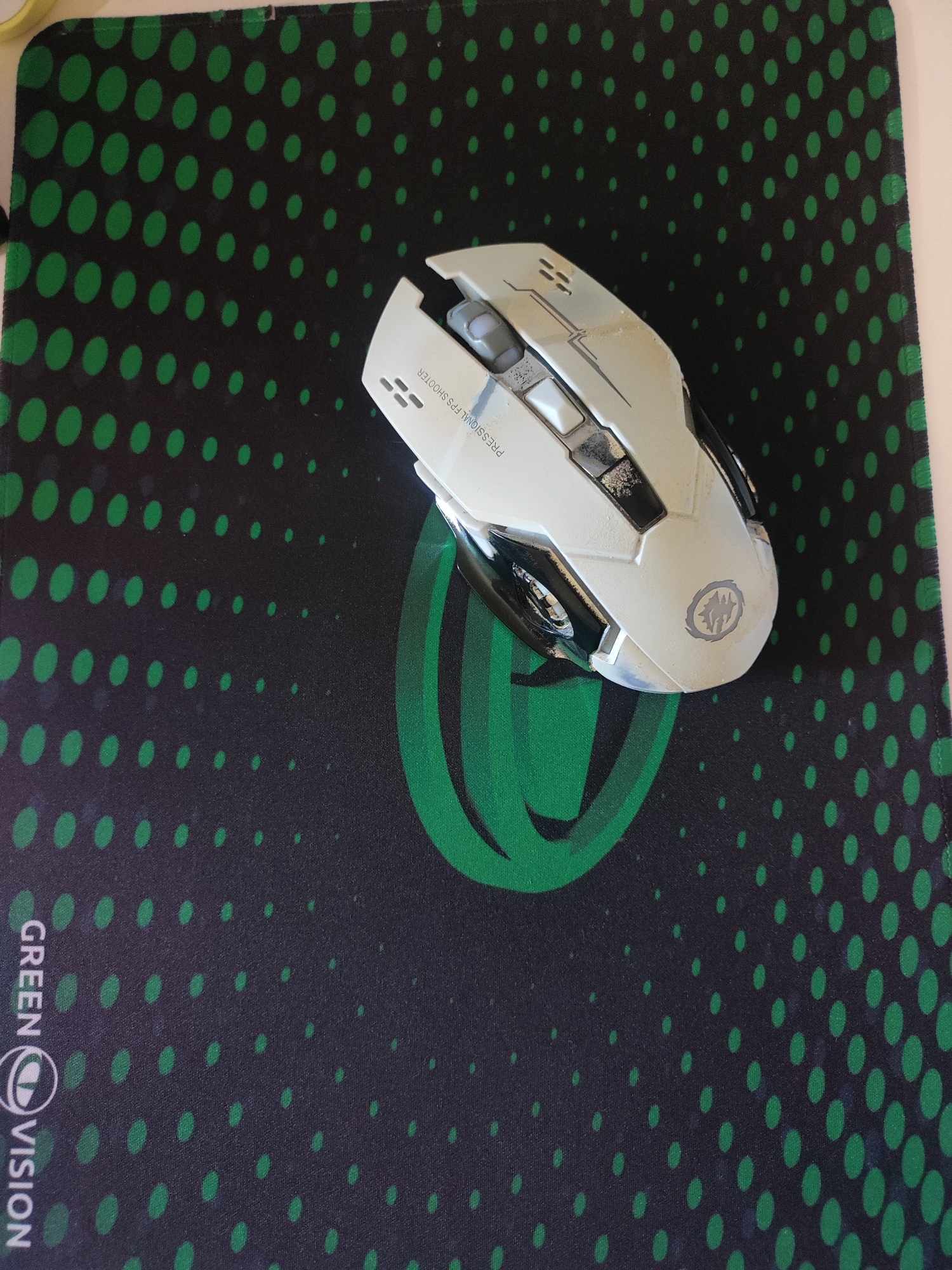 The image shows a white and silver gaming mouse placed on a black mousepad with green circular polka dots. The mousepad has a green stylized "G" logo and the text "GREEN VISION" printed vertically along the bottom left edge. The mouse has visible wear and tear, suggesting it has been used. The text "PROFESSIONAL FPS SHOOTER" is printed on the side of the mouse. The overall setting appears to be an indoor environment, likely a desk. There is no information in the image to suggest the location is the city of Cherkasy, Ukraine.