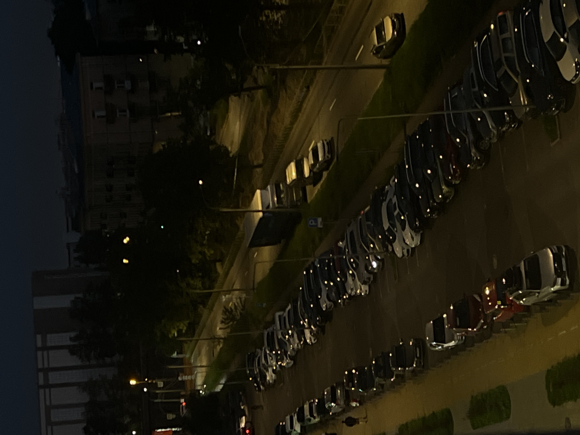 The image shows a nighttime aerial view of a street in Saint Petersburg, Russia, with a parking lot filled with cars. The street is lined with trees and lampposts, and there are buildings visible in the background. The cars are parked parallel to the street, and many of them have their headlights on. A few people are visible walking on the sidewalk next to the parking lot. The scene is dimly lit by streetlights and the lights from the cars and buildings.