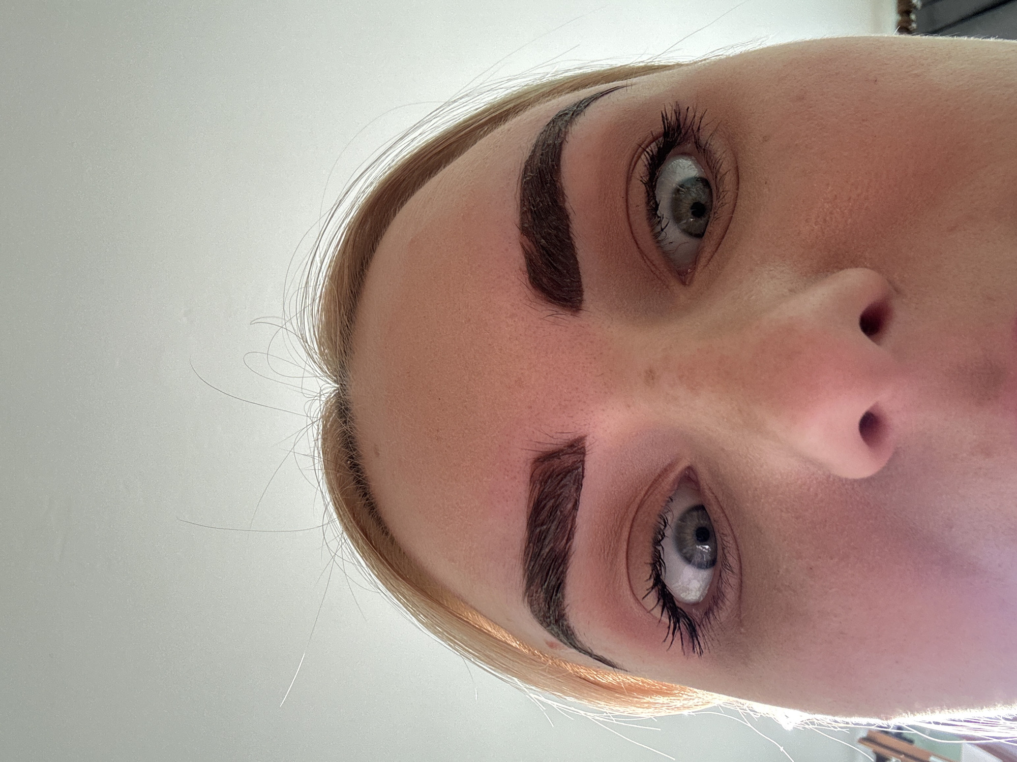 The image is a close-up, low-angle shot of a person's face, focusing on their eyes and forehead. The subject has blonde hair and is looking upwards with wide, surprised eyes. Their eyebrows are dark and sharply defined. The lighting suggests an indoor setting with a light source from above. The background is a plain, light-colored surface, likely a ceiling. No specific location in the city of Unknown, Russia can be determined from the image.