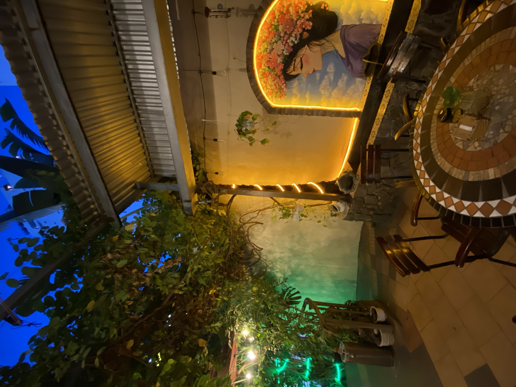The image depicts an outdoor seating area of a restaurant or cafe in Nha Trang, Vietnam, likely in the evening given the artificial lighting. The scene is characterized by lush greenery, with vines and plants draping from an overhead structure. In the foreground, a circular mosaic-topped table is surrounded by dark wooden chairs.
To the right, a wall features a mural of a woman wearing a floral crown, framed by a glowing orange neon outline. The wall behind the mural is painted a pale yellow and appears to be lit by ambient light. Further to the left, green LED lights illuminate a section of the foliage and a wooden railing. A tall, cylindrical trash bin and some potted plants are visible near the railing.
The overhead structure is partially covered with corrugated metal roofing, suggesting it's a covered patio. The overall ambiance is one of relaxed outdoor dining, with a bohemian or tropical aesthetic. The sky above is a deep blue, indicating twilight or early night. The flooring is paved with square tiles in various shades of brown and terracotta.