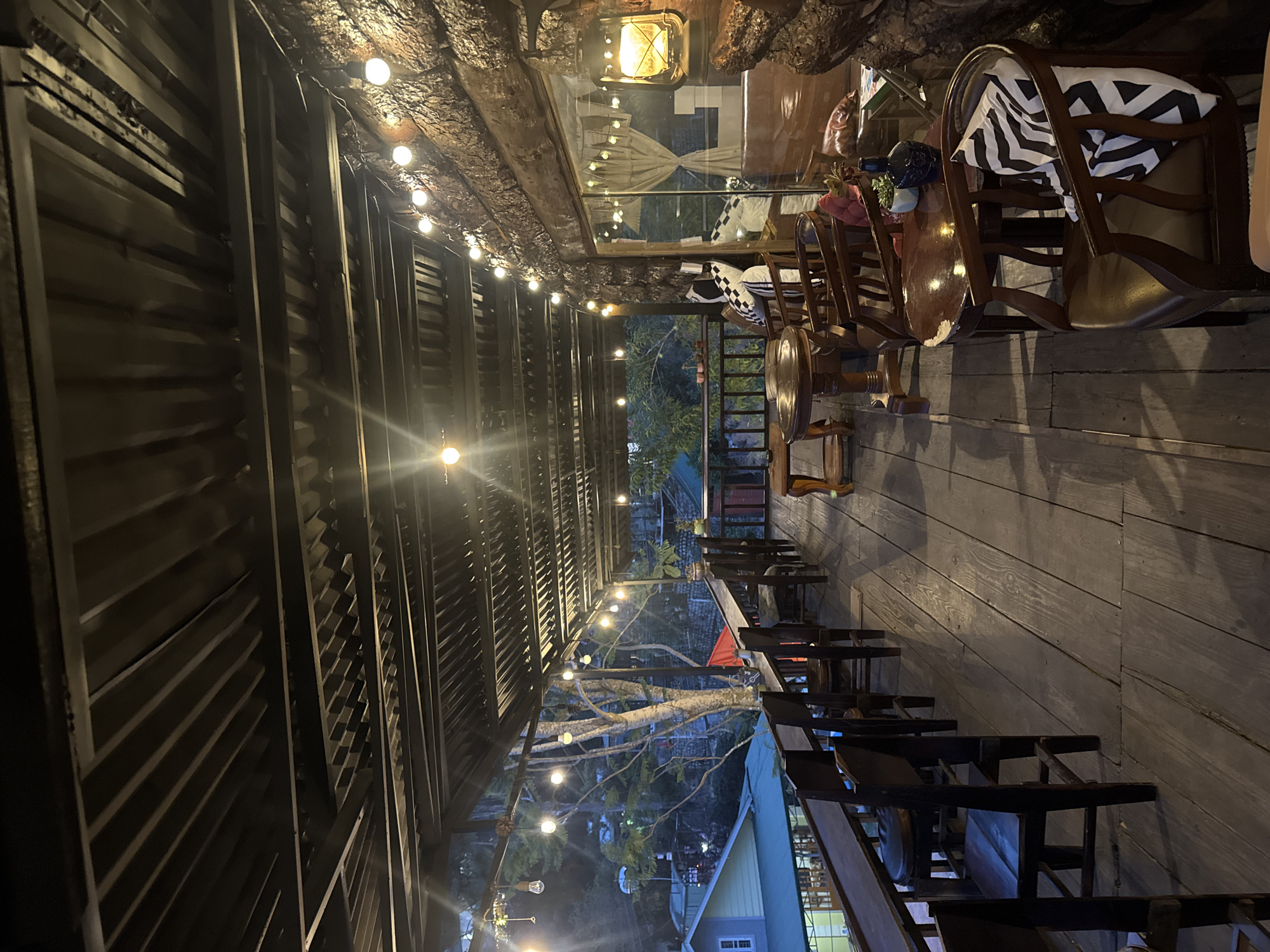 The image depicts an outdoor restaurant or cafe, likely in Xuân Trường, Đà Lạt, Vietnam, based on architectural cues and possible lighting. The scene is captured at night, lit by a series of string lights strung along the ceiling and a lantern hanging near the entrance. The ceiling is constructed from rough, possibly natural, materials like wood and stone. The left side of the scene shows a structure with a corrugated metal roof or wall, further reinforced by metal supports. The right side features wooden flooring and tables arranged along the length of the space. The chairs are primarily made of wood, some with cushions, including one with a black-and-white striped pattern. Tables are set with decorations such as plants and vases. Beyond the covered area, trees and buildings are visible, suggesting an open-air aspect to the venue.