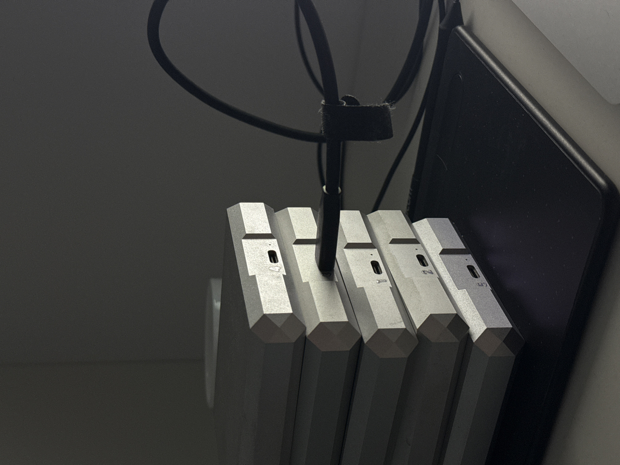 An indoor, low-light photograph captures a collection of electronic devices arranged vertically. Five silver-colored, hexagonal prism-shaped objects, possibly external hard drives or small form-factor computers, are stacked or lined up. A black power or data cable descends from above, secured by a black hook-and-loop strap, and connects to one of the units. Other black cables are visible in the dimly lit background. To the right, a dark, rectangular electronic device, likely a tablet, leans against a lighter surface. Each hexagonal unit has a small rectangular port on its top surface, accompanied by a numeric label. From left to right, the clearly visible labels on the units are '4', '3' (the unit into which the cable is plugged), '2', and '5'. The overall scene suggests an organized setup of electronic equipment in a workspace or server area, under low ambient lighting. The location is Башкултаево, Russia.