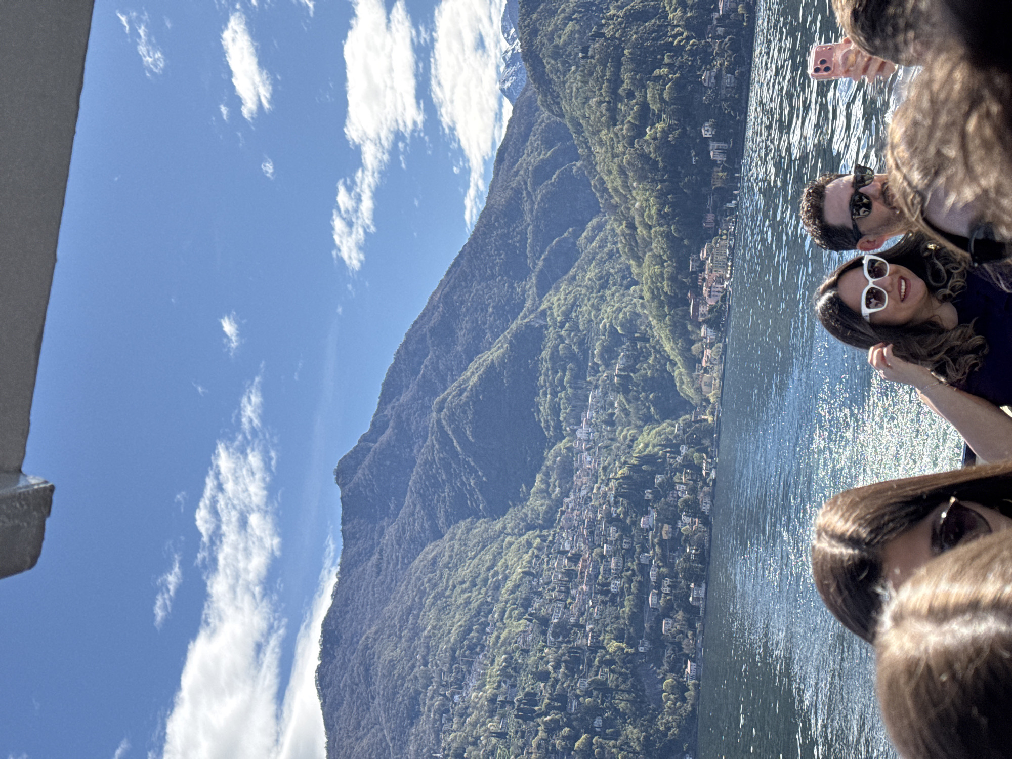 The image shows a view of the mountainous landscape of San Siro, Italy, with Lake Como in the foreground. A group of people, including a man and a woman with sunglasses, are present on what appears to be a boat or platform, taking photos. The town of San Siro is situated on the mountainside. The sky is blue with scattered clouds.