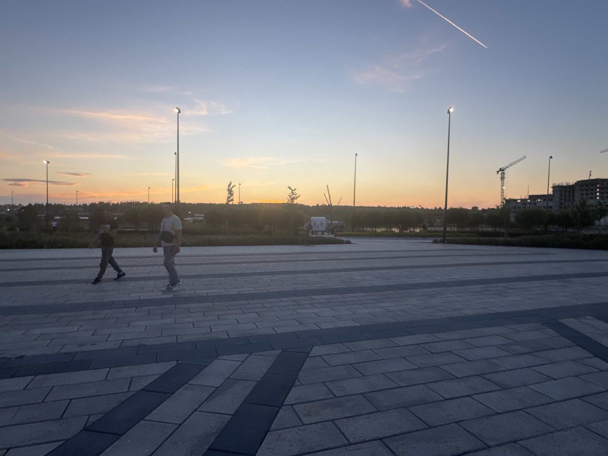 This photograph depicts an outdoor urban scene at dusk, with a clear sky transitioning from a pale orange near the horizon to a deep blue overhead. A contrail from an aircraft is visible high in the sky. In the middle ground, two individuals are walking across a paved plaza composed of rectangular gray and darker gray stones. The person on the left is wearing dark pants and a dark shirt, while the person on the right is in lighter pants and a light shirt, carrying a bag over their shoulder. Several tall, slender lampposts are scattered across the scene, some illuminated with a warm light. A line of trees forms a dark barrier in the background, behind which are glimpses of construction cranes and unfinished buildings, suggesting ongoing development. The overall impression is of a modern, developing area in a city. Based on the context provided, this scene is located in the city of Magnitogorsk, Russia.