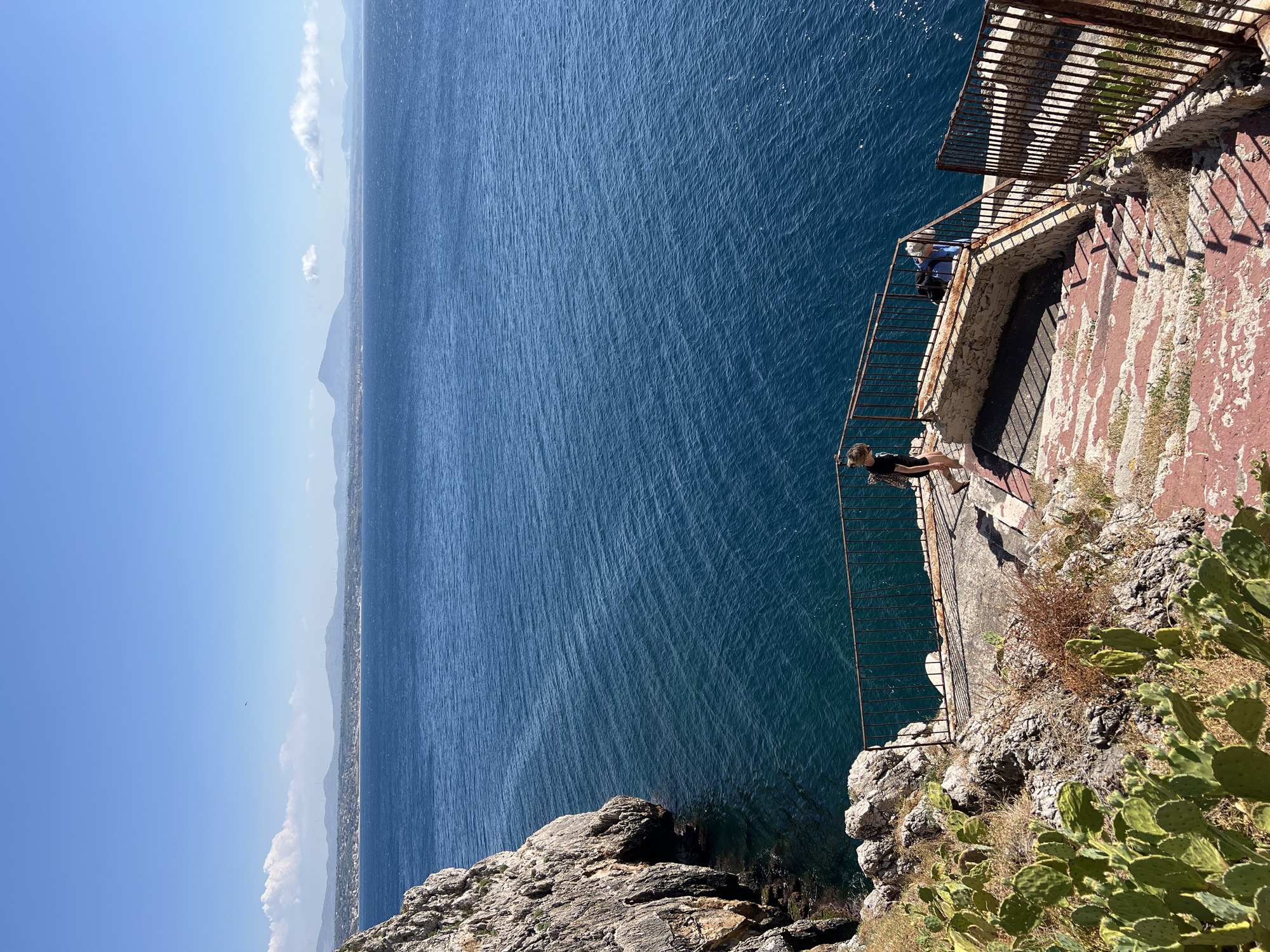 The image shows a scenic view of the sea from a cliffside. A woman is standing on a narrow walkway overlooking the vast blue ocean. The walkway is made of stone and has a rusty metal railing. There are also some prickly pear cacti growing on the cliff. In the background, a distant coastline with mountains can be seen. The sky is clear and blue, suggesting a sunny day. The sun's glare on the water indicates it is likely midday. The overall scene evokes a sense of peace and tranquility, with the woman appearing to be enjoying the view. The location appears to be Terrasini, Italy, known for its beautiful coastal scenery.