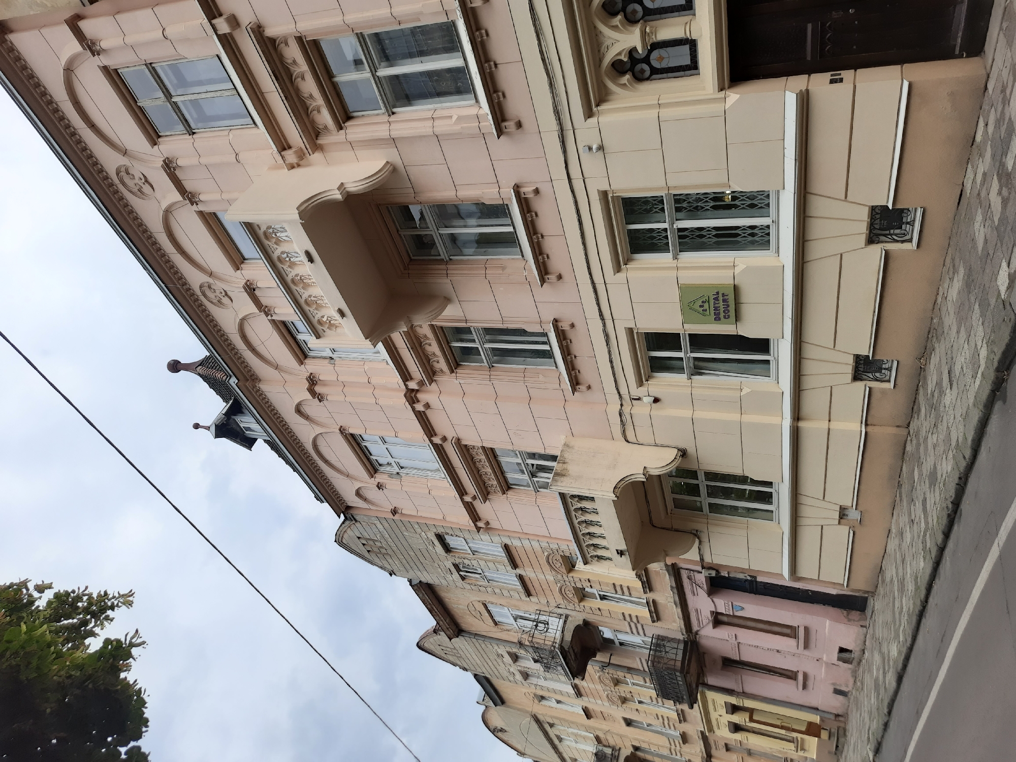 The image shows a street scene in Lviv, Ukraine, on a cloudy day. The perspective is from street level, looking up at the facades of several buildings. The buildings are of varying heights and architectural styles, predominantly featuring a pastel pink and beige color palette. Decorative elements such as balconies, ornate window frames, and arched detailing are visible.
On the right side of the image, a building with a cream-colored facade has a green rectangular sign that reads "DENTAL COURT". Adjacent to this sign is a large dark wooden door with carved detailing. Below the windows, small decorative grates are installed.
Further to the left, the buildings transition to a lighter pink and then to weathered stone and plaster facades with visible signs of age. Several balconies with wrought-iron railings are present on these older structures. The street in the foreground is paved with cobblestones, and a white line marks the edge of the road. Overhead, tree branches with green leaves are visible on the left, and power lines crisscross the sky. The overall atmosphere suggests an older, historic urban environment.