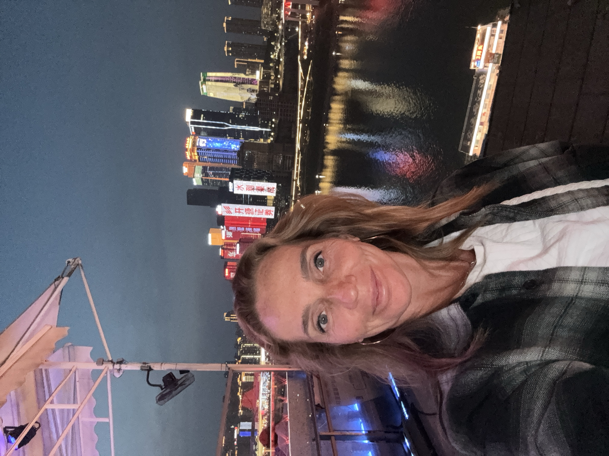 A woman takes a selfie at night in Yuzhong District, China, with a background of lit buildings and a river. The city lights are reflected in the water. She is wearing a white shirt and plaid jacket, with gold hoop earrings. A structure with lights is in the lower left.